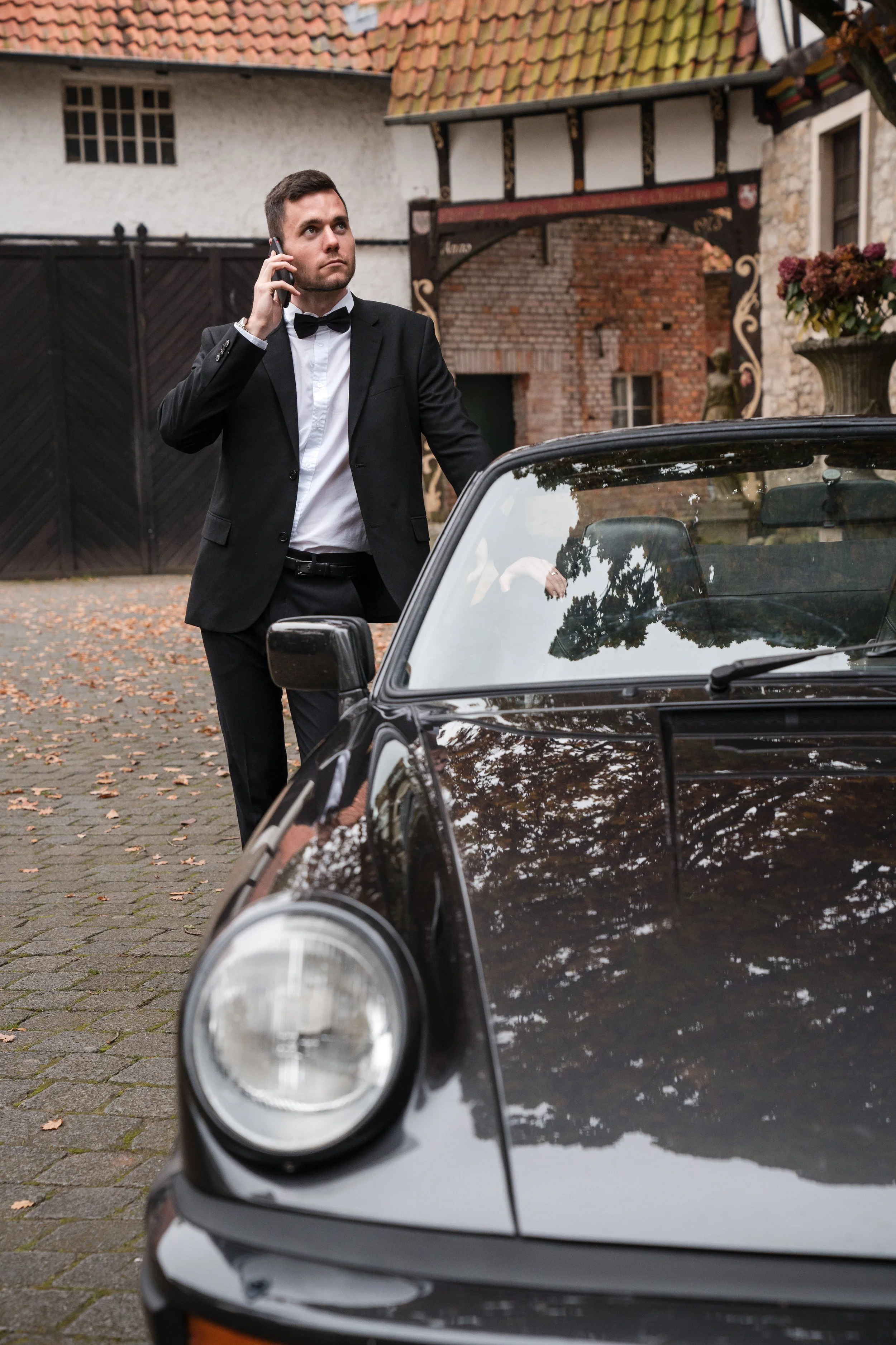 Bräutigam in einem schwarzen Anzug mit Fliege steht neben einem schwarzen Sportwagen im Freien, während er mit einem Mobiltelefon spricht.