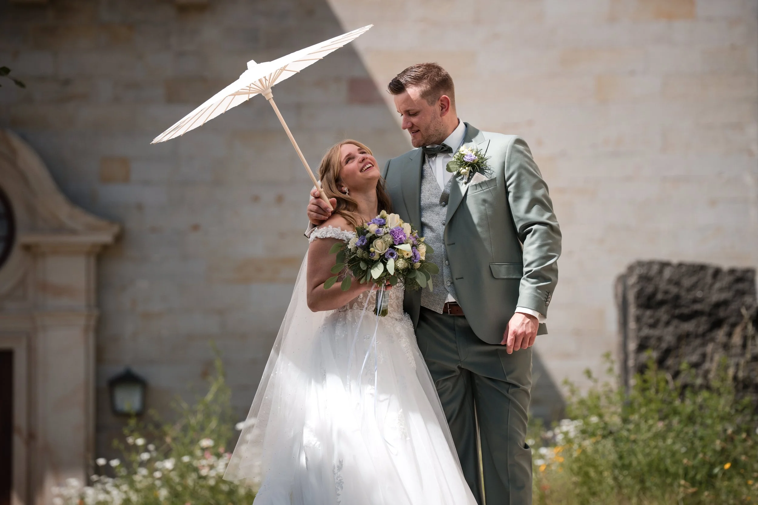 Ein glückliches Brautpaar bei einer Hochzeit im Freien, die Braut hält einen Blumenstrauß und beide lächeln sich an, während der Bräutigam sie mit einem weißen Sonnenparasol schützt.