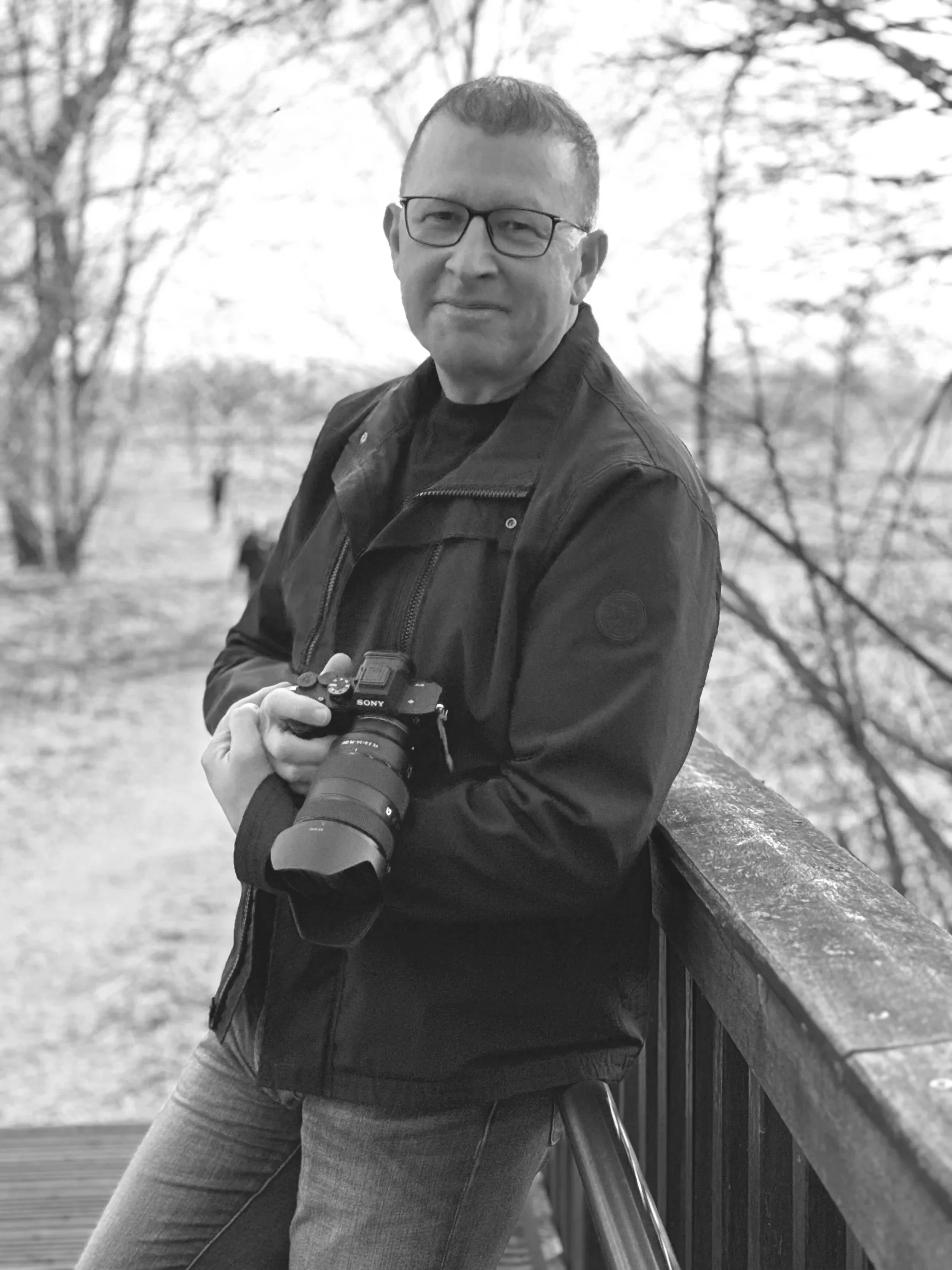 Ein Mann mit Brille trägt eine schwarze Jacke und hält eine Kamera in der Hand, steht an einem Geländer im Freien mit Bäumen im Hintergrund.