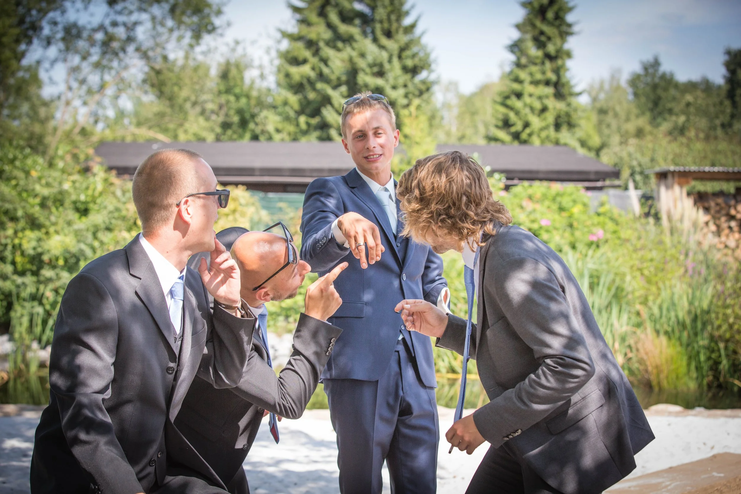 Vier Personen in Anzügen, die im Garten miteinander reden und lachen. Eine Person zeigt auf eine andere. Im Hintergrund Bäume und ein Holzgebäude.