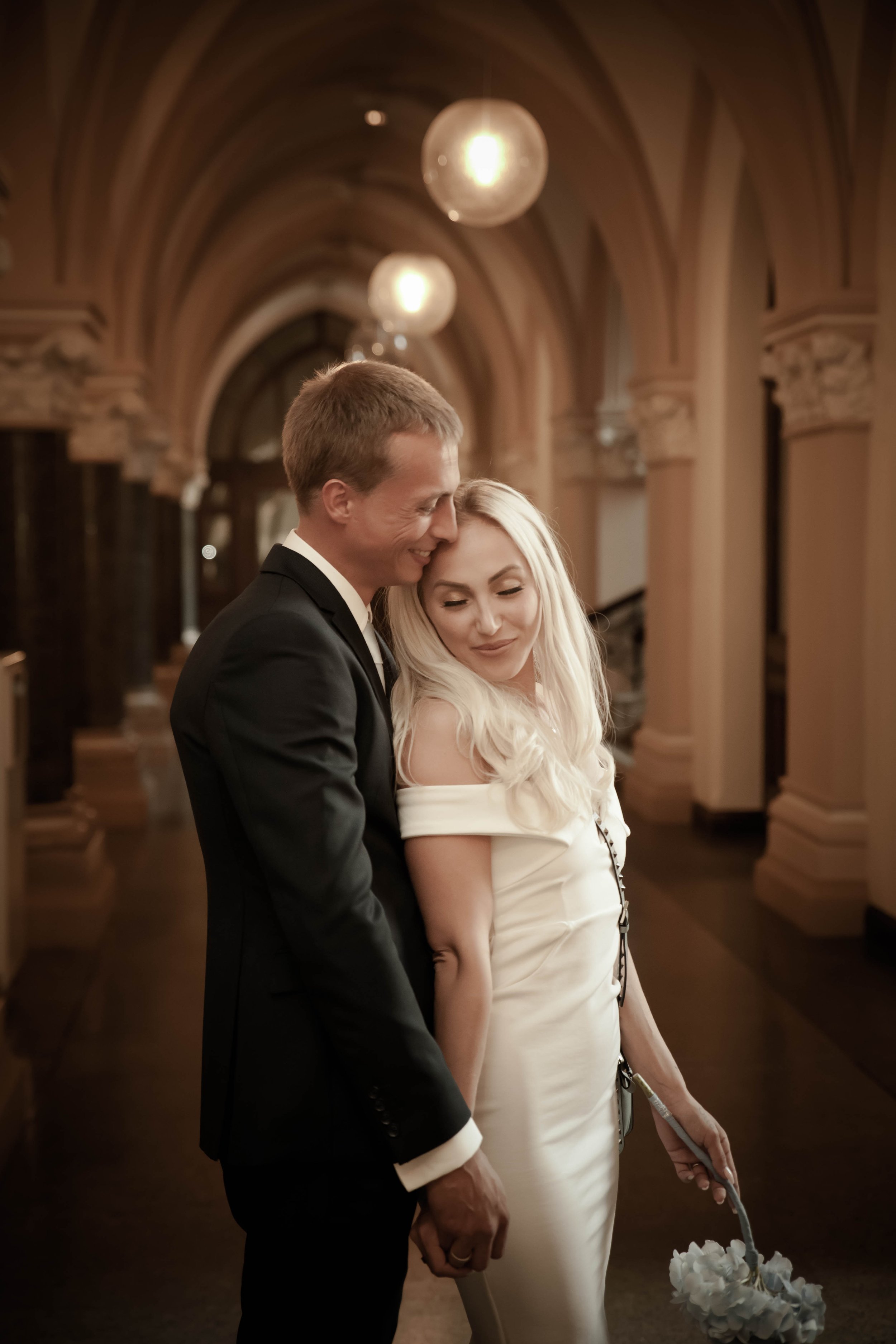 Ein verheiratetes Paar bei ihrer Hochzeit in einem eleganten Saal, Hand in Hand, die Braut trägt ein weißes Kleid mit Dekoration, der Bräutigam trägt einen schwarzen Anzug. Sie hinterlassen einen liebevollen Eindruck.