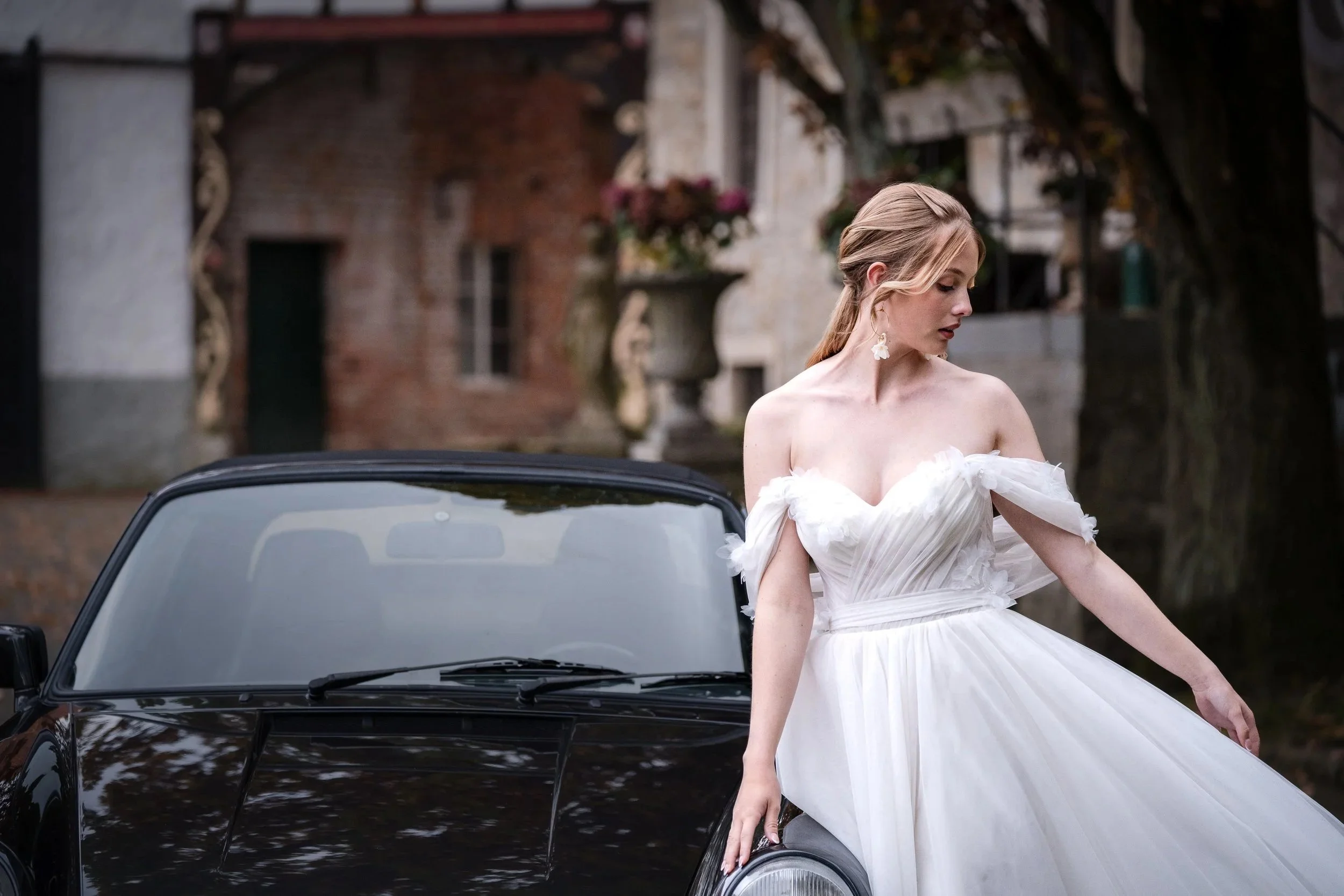 Eine junge Braut in einem weißen Brautkleid steht neben einem schwarzen Porsche, in einer malerischen, historischen Hochzeit Location Umgebung mit alten Gebäuden und Bäumen in Hannover