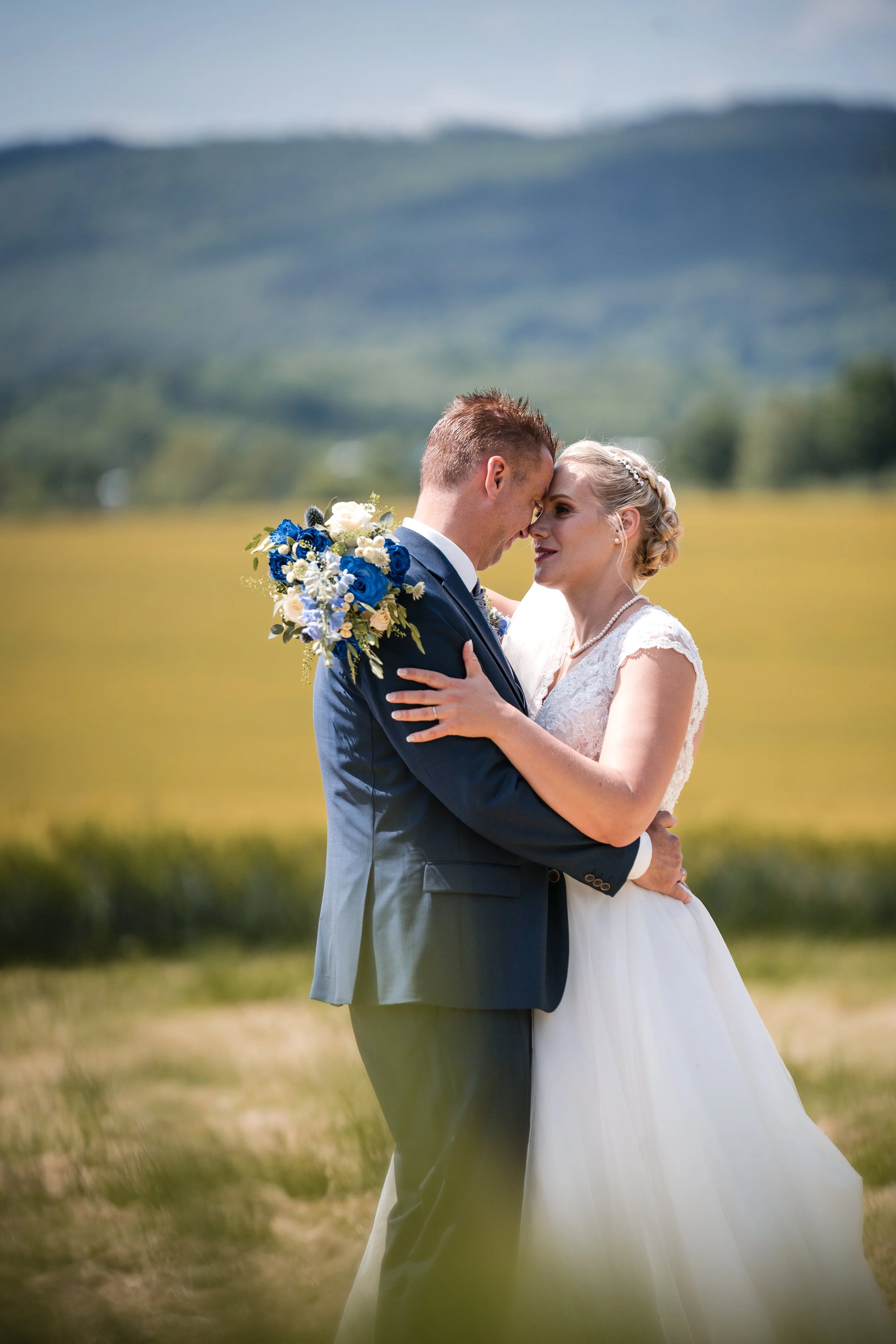 Ein Brautpaar bei einer Hochzeit im Freien, umarmend, mit Bergen und Himmel im Hintergrund.