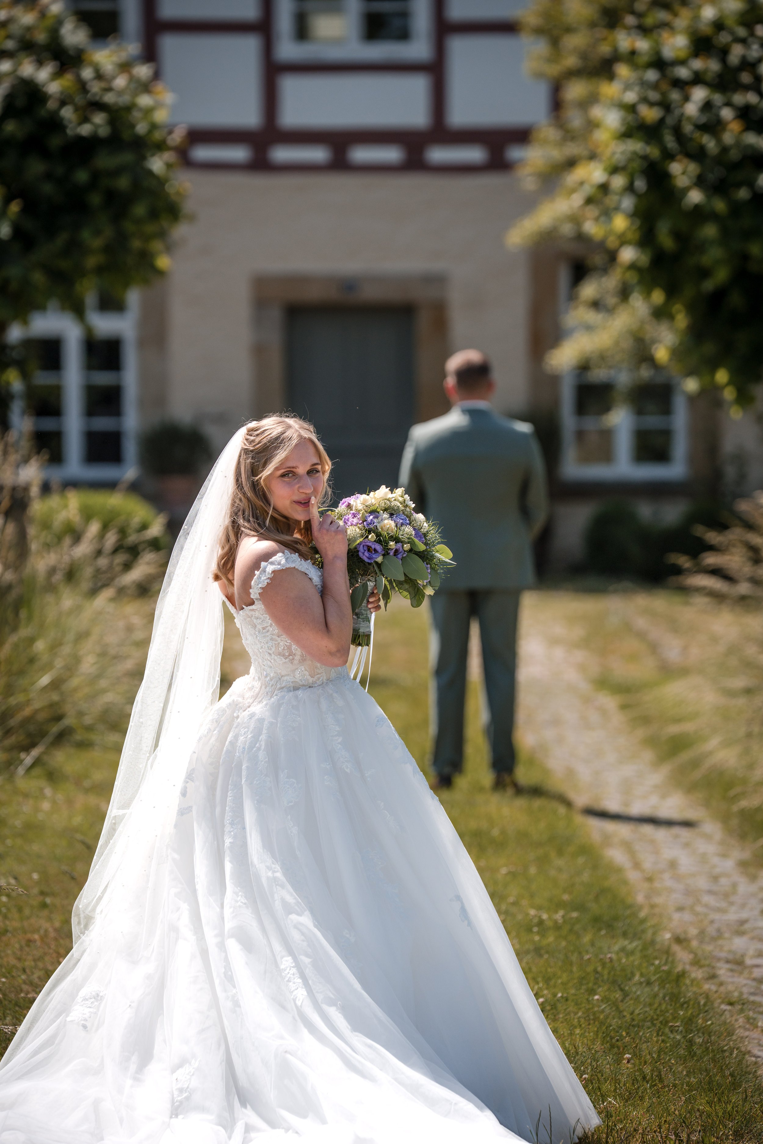 Eine Braut trägt ein weißes Brautkleid mit Schleier und hält einen kleinen Blumenstrauß, während sie vor einem Haus steht. Im Hintergrund ist ein Mann in Anzug, der von hinten sichtbar ist.