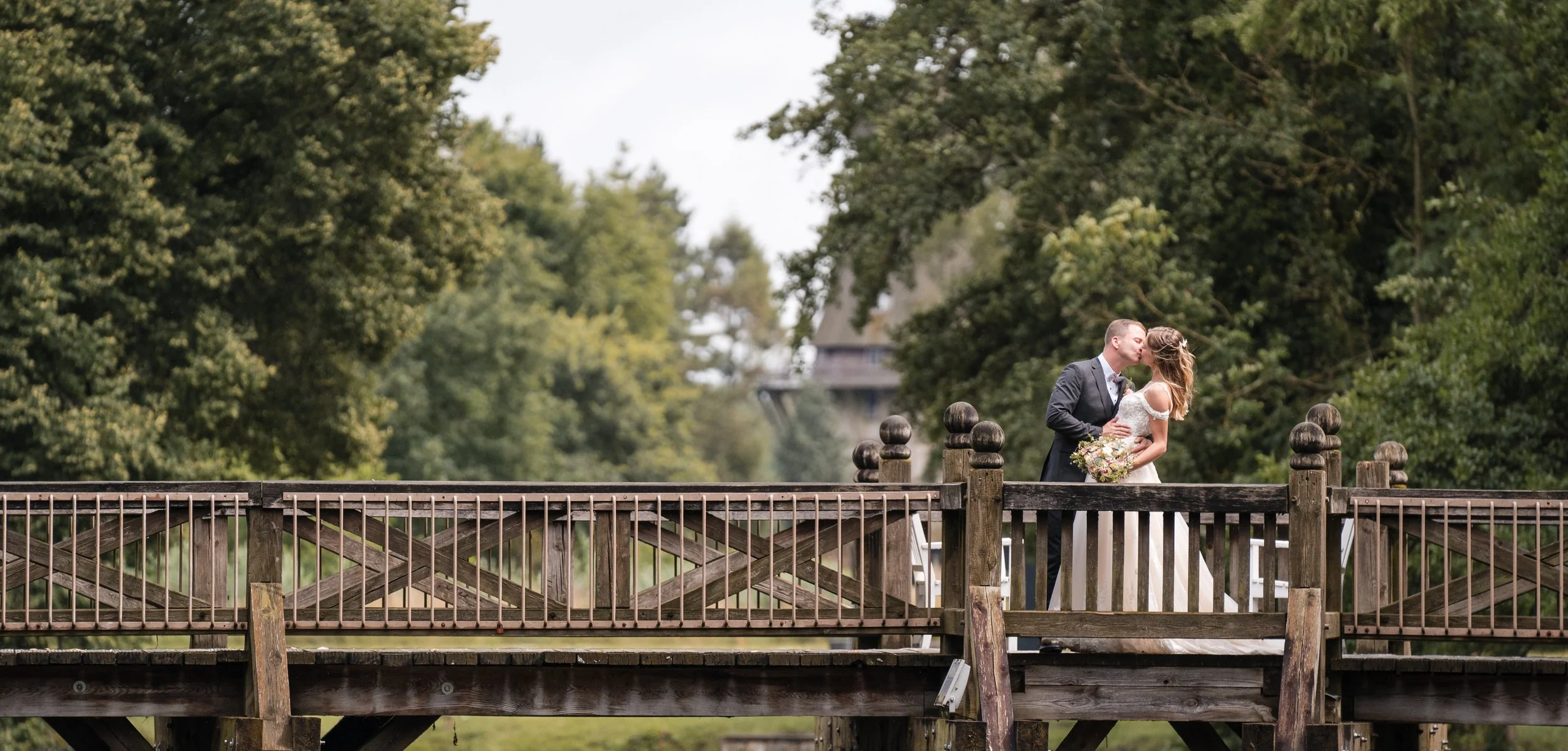 Ein Brautpaar küsst sich auf einer Holzbrücke, umgeben von grünen Bäumen in Gifhorn.
