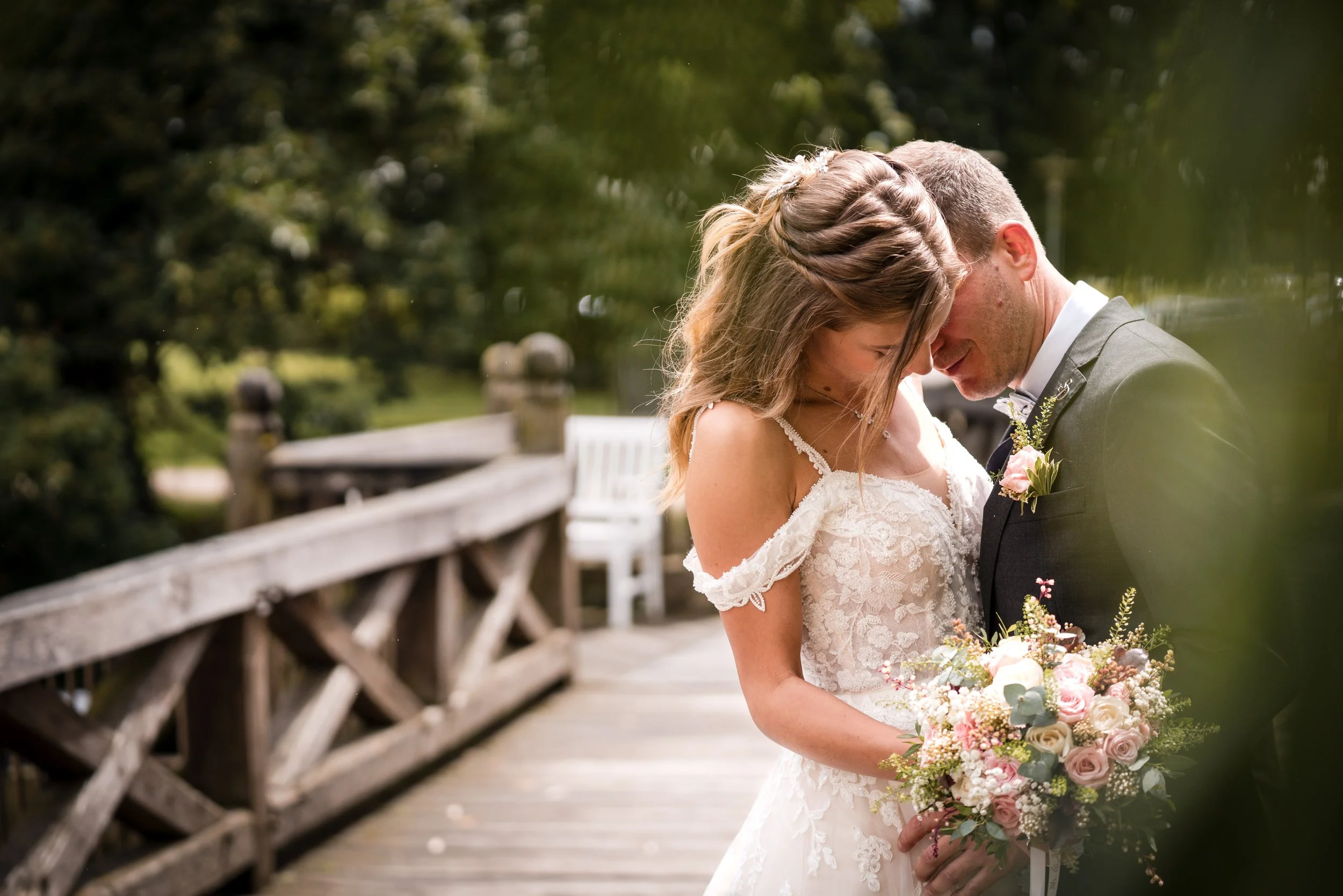 Ein Brautpaar steht sich an einem bewaldeten Ort gegenüber und schaut liebevoll auf die Hochzeitsblumen. Die Braut trägt ein weißes Spitzenkleid, der Bräutigam einen dunklen Anzug, beide mit kleinen Blumen im Revers. Im Hintergrund eine Holzbrücke und grüne Bäume.