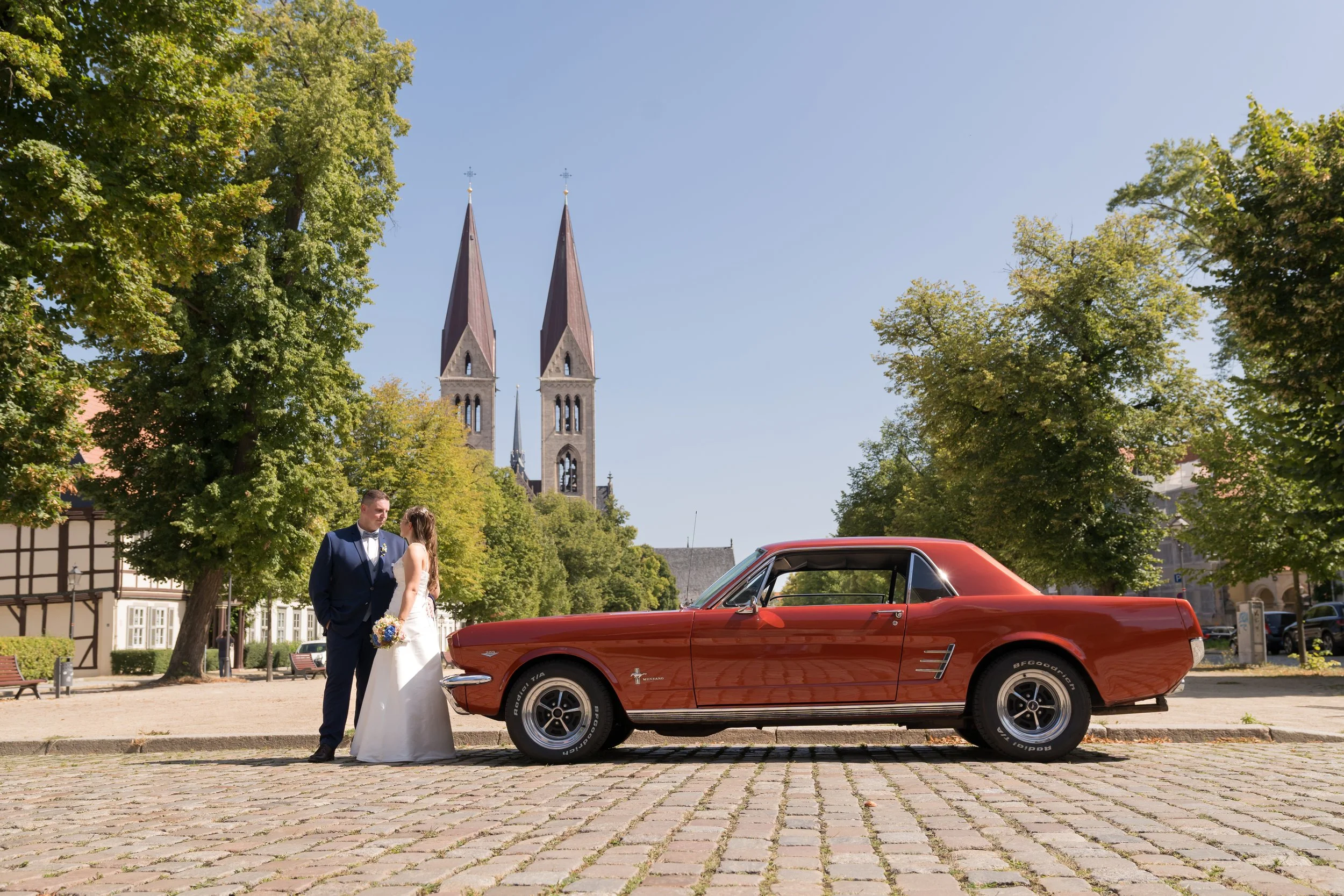 Ein Brautpaar steht neben einem roten Oldtimer in einer Stadt mit Kirche im Hintergrund, sonniger Tag.