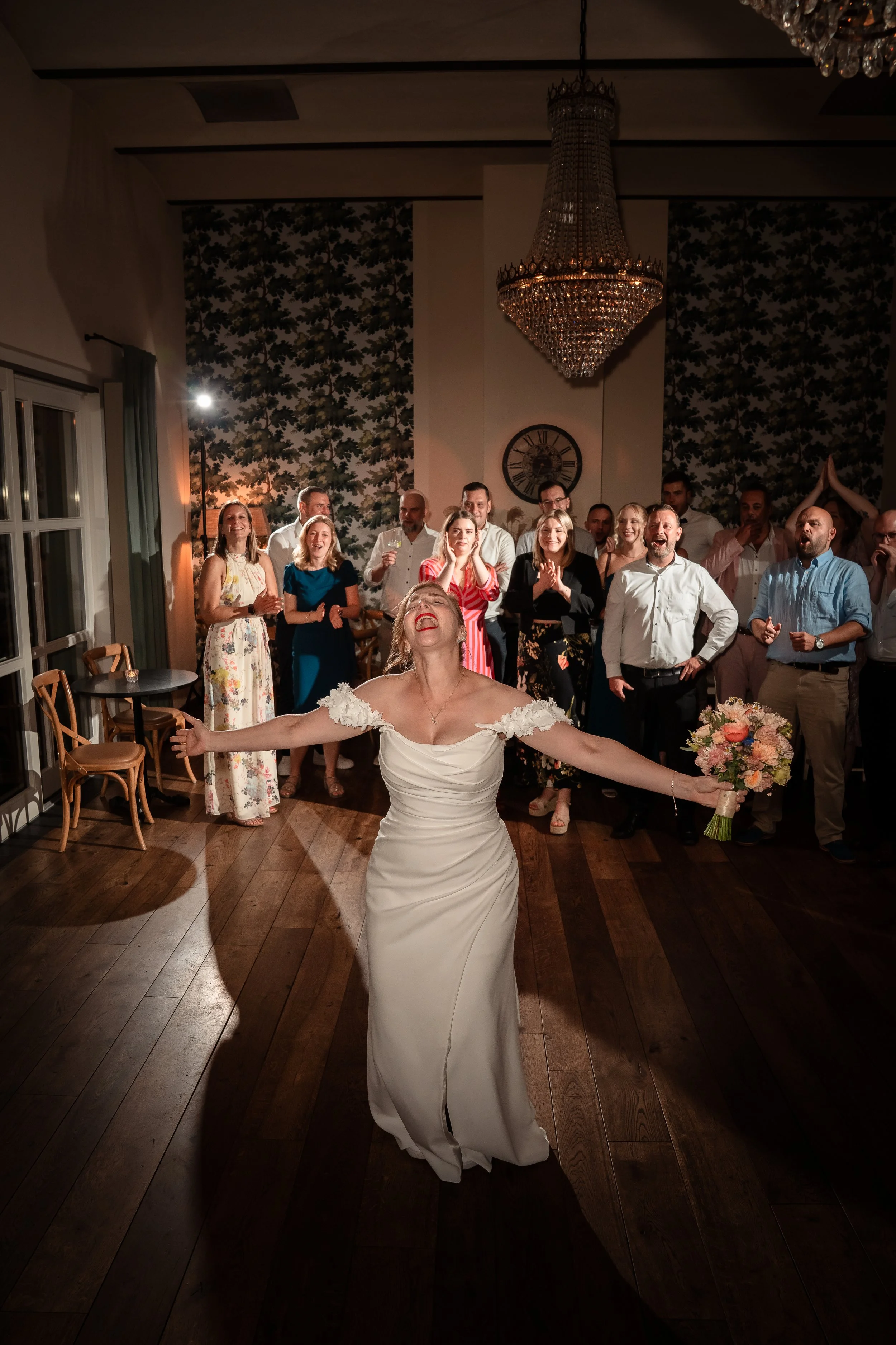 Eine braut in einem weißen Kleid steht im Mittelpunkt, mit Armen offen, während sie vor einer Gruppe von lachen Menschen kurz vor ein Brautstraußwurf bei einer Hochzeit im festlichen Raum steht.