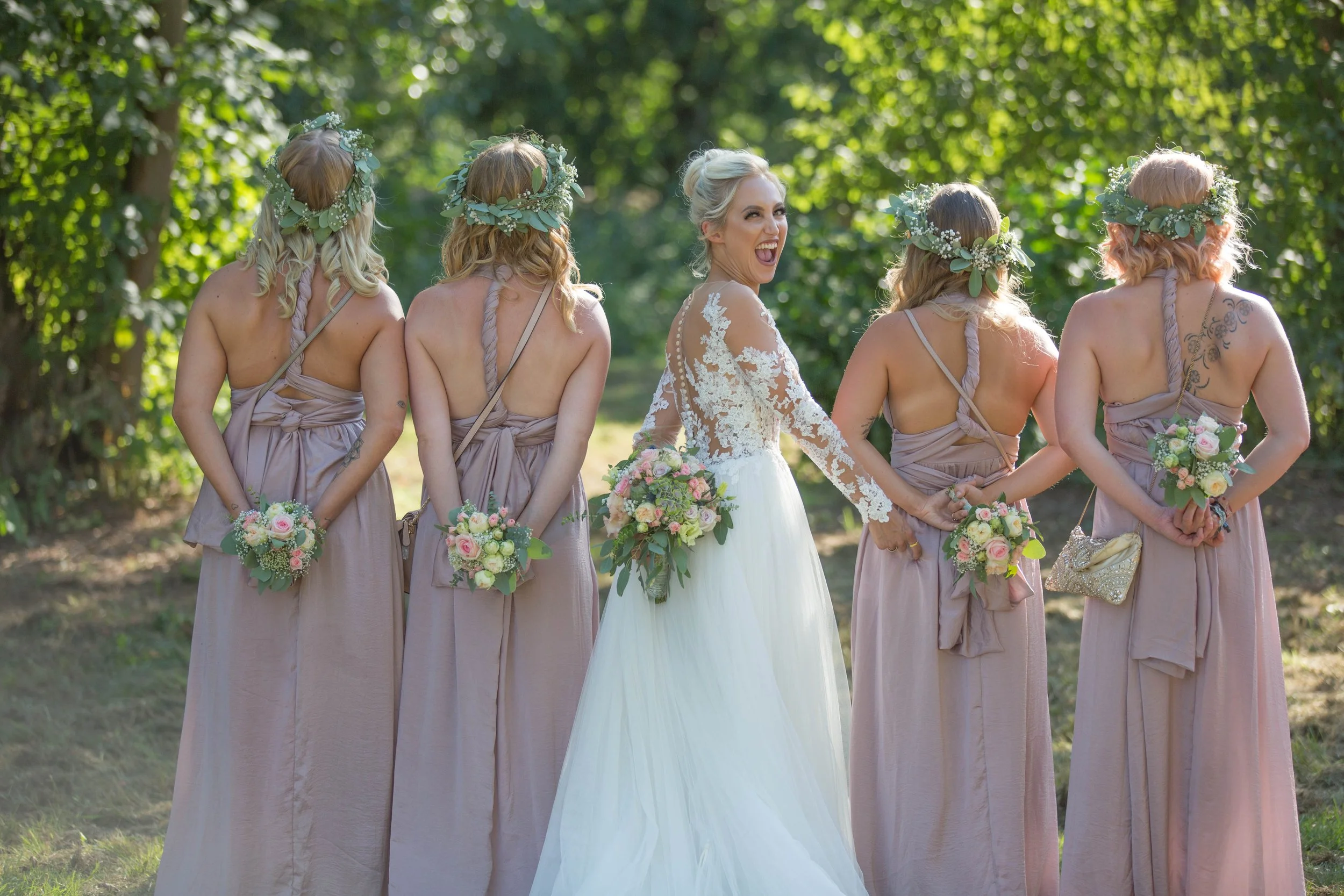 Braut und vier Brautjungfern im Garten, lachend, mit Blumenkränzen und Blumensträußen, bei einer Hochzeit im Freien.