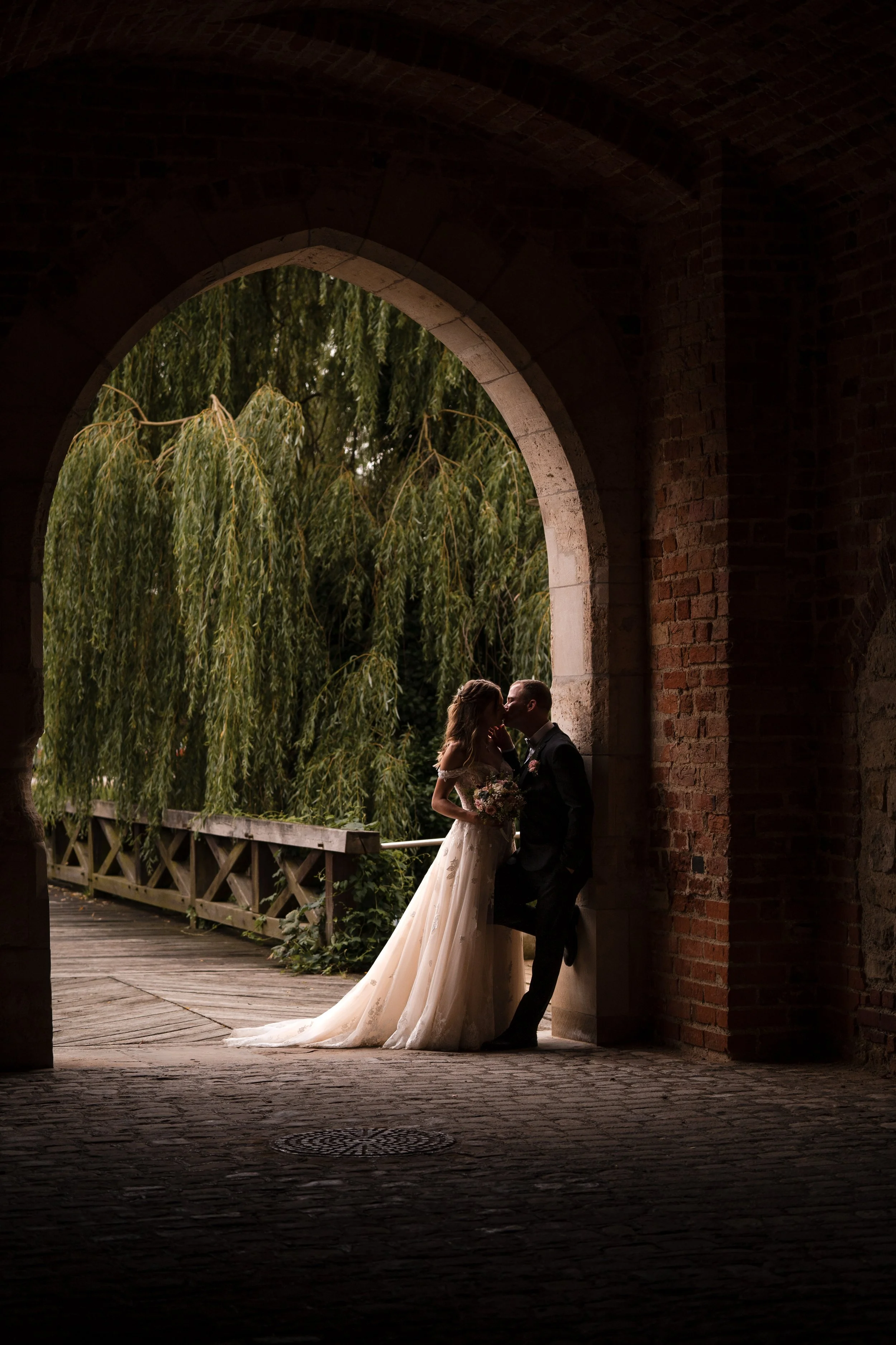 Ein Brautpaar küsst sich unter einem Bogen in einer alten Burg, während im Hintergrund ein Baum mit herabhängenden Ästen steht in Deutschland.