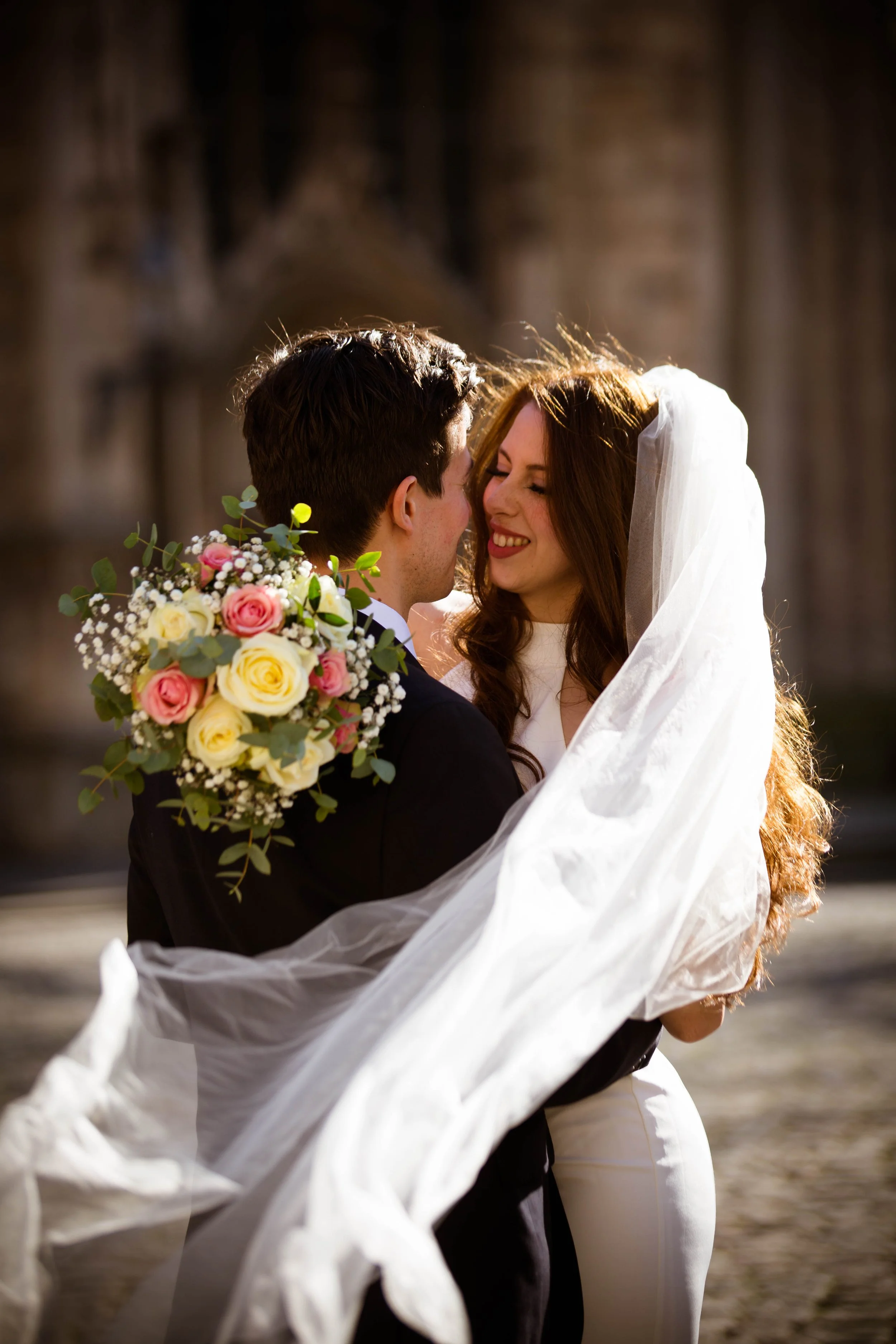 Ein Brautpaar umarmt sich lachend, die Braut trägt einen Schleier, der im Wind weht, und hält einen Blumenstrauß aus weißen und rosa Rosen. Hintergrund ist eine alte Kirche im Sonnenlicht.