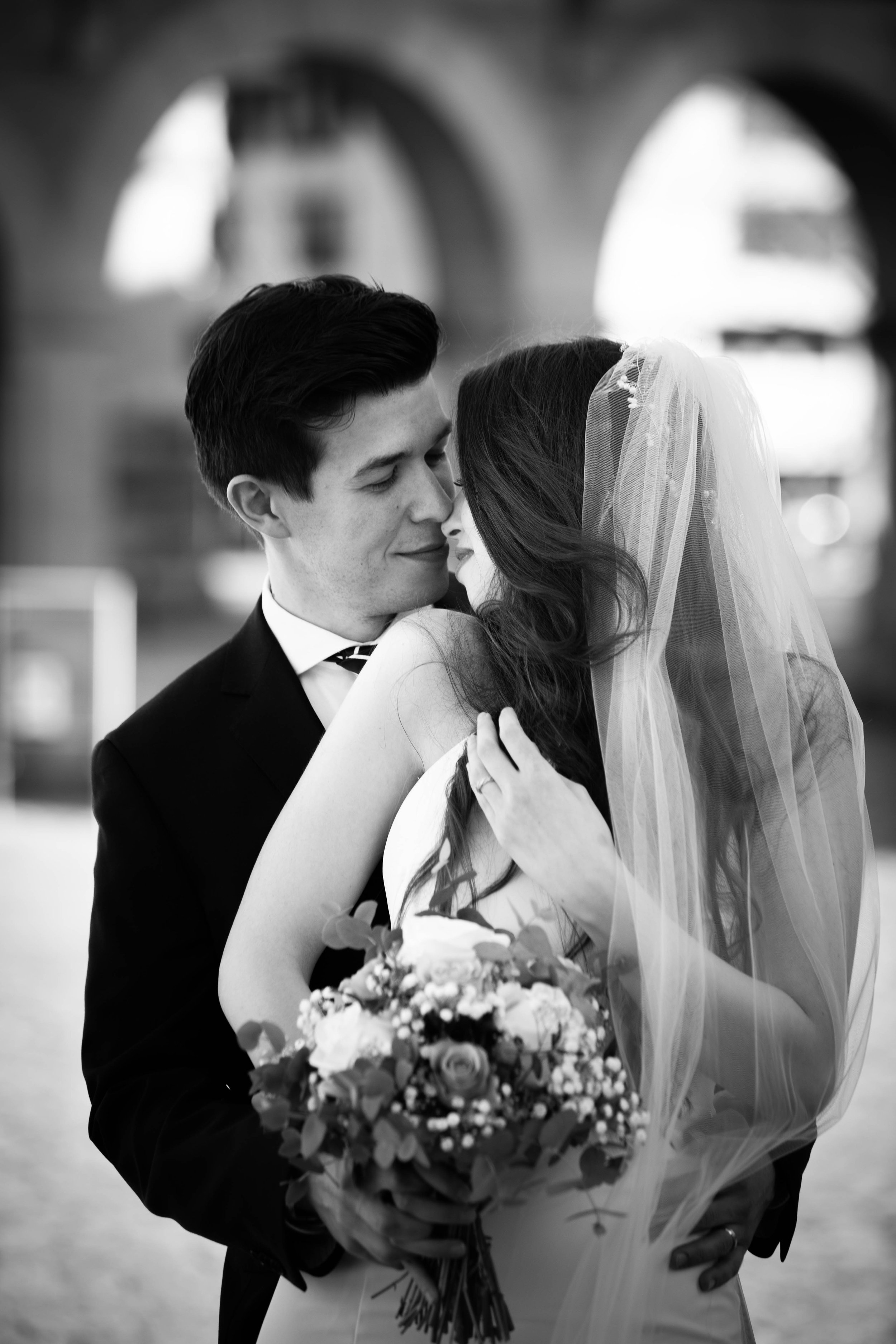 Ein Brautpaar in Hochzeitstracht, umarmend, mit Brautstrauß, bei einer emotionalen Umarmung, in Schwarz-Weiß-Fotografie.