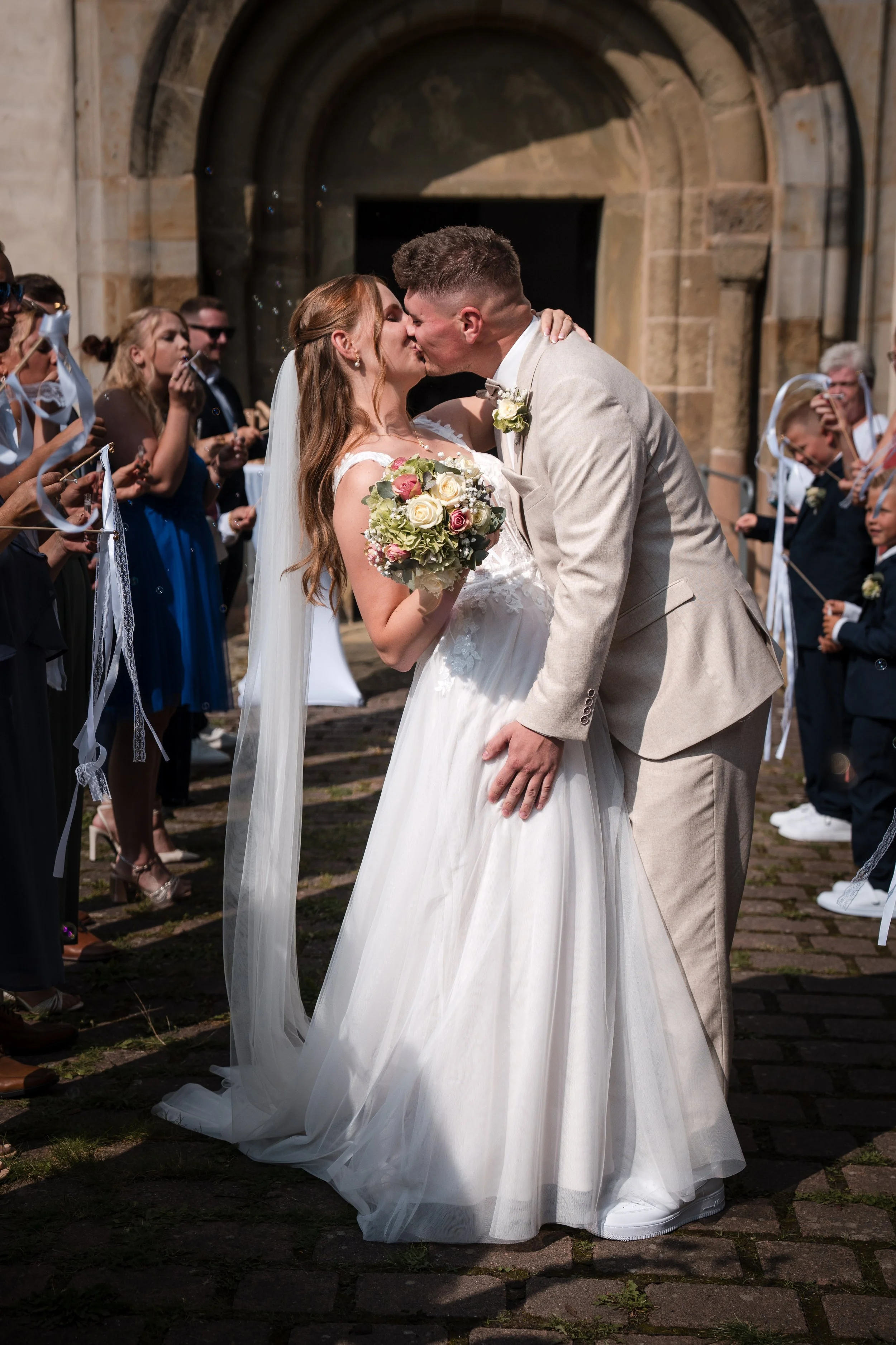 Ein Brautpaar küsst sich während ihrer Hochzeit im Freien, umgeben von Gästen, vor einem historischen Gebäude.