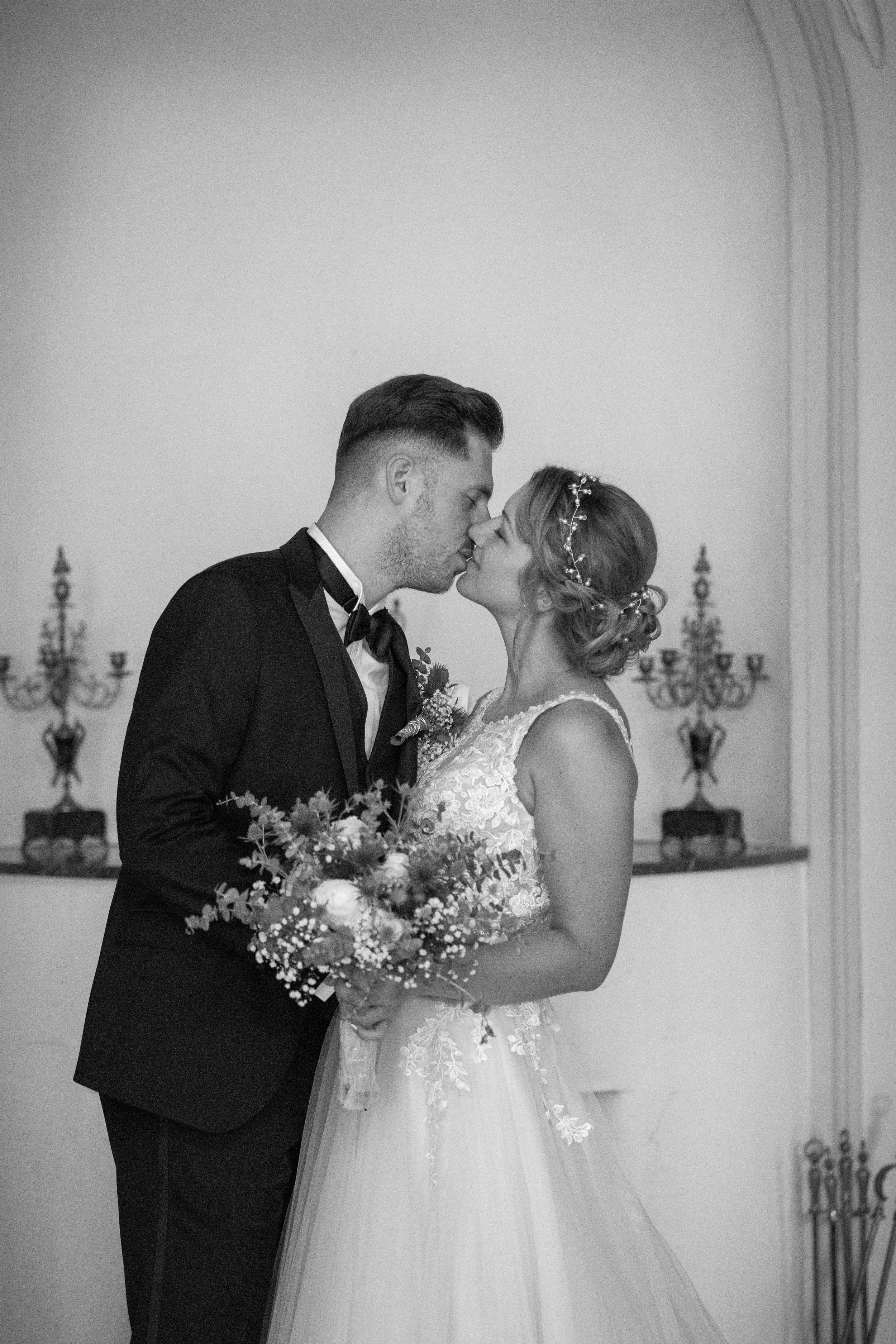 Braut und Bräutigam küssen sich bei ihrer Hochzeit, in eleganten Kleidungsstücken, im Hintergrund Wandhalterungen.