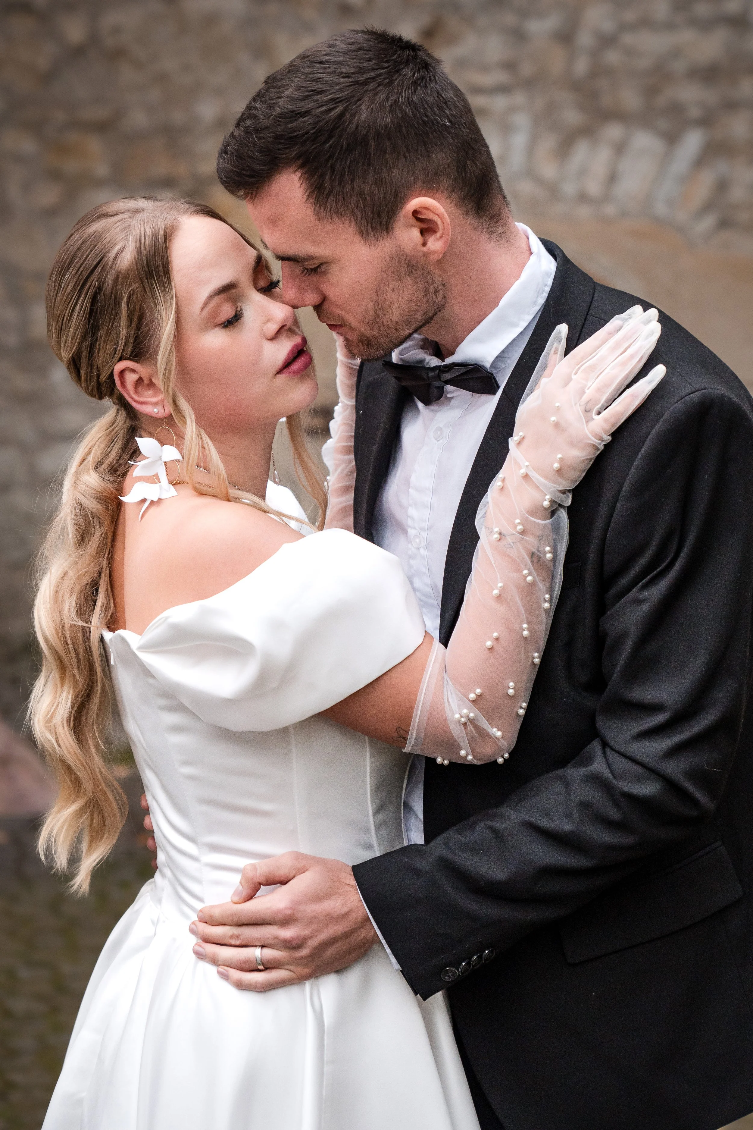 Ein Paar in Hochzeitskleidung tanzt eng umeinander, die Frau trägt ein weißes Hochzeitskleid mit Glitzerarmbanden und der Mann einen schwarzen Anzug mit Fliege, im Hintergrund eine Steinmauer.