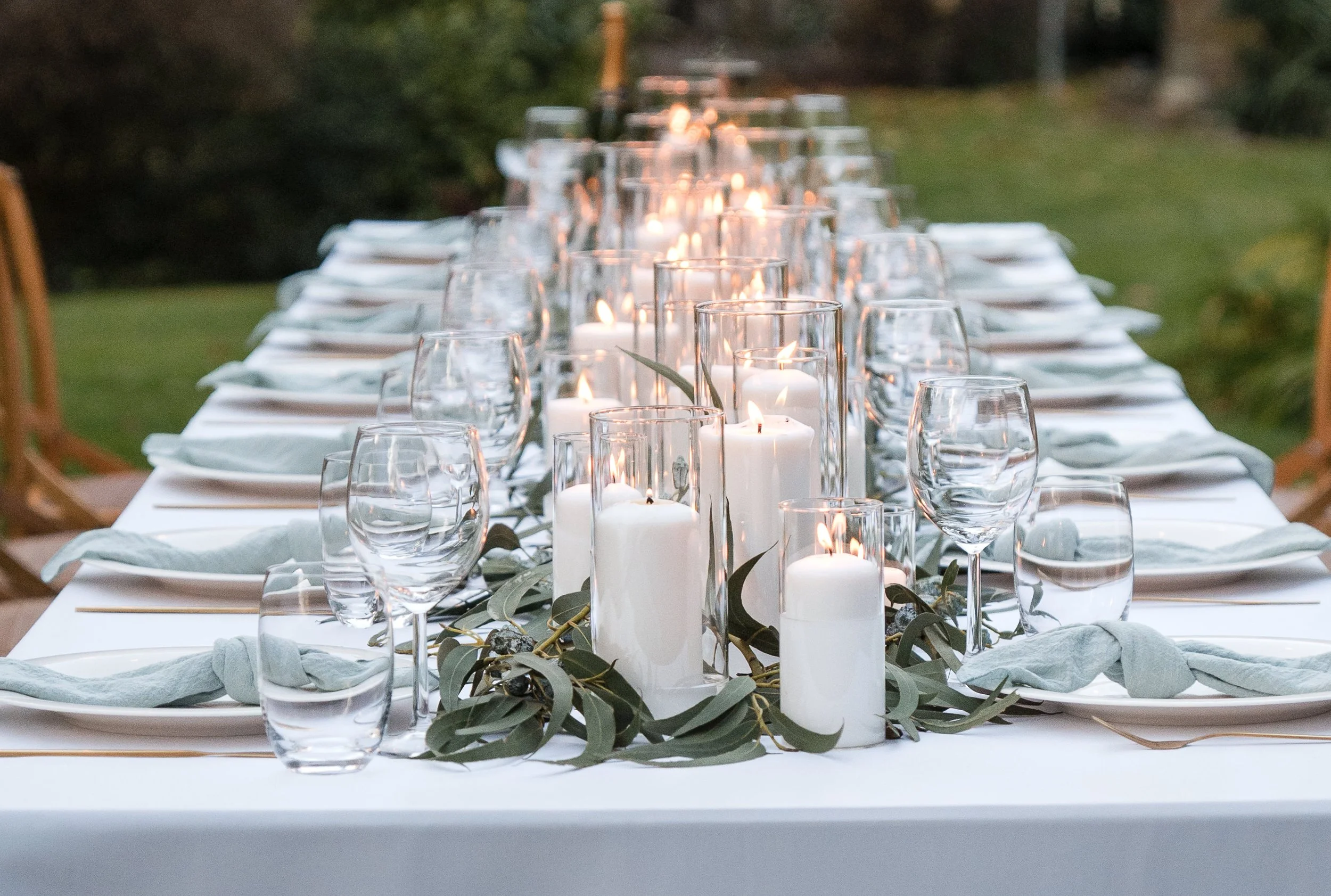Ein eleganter Esstisch im Freien, dekoriert mit weißen Kerzen in Glasvasen und frischem grünem Laub, für eine festliche Hochzeit in der Toskana..