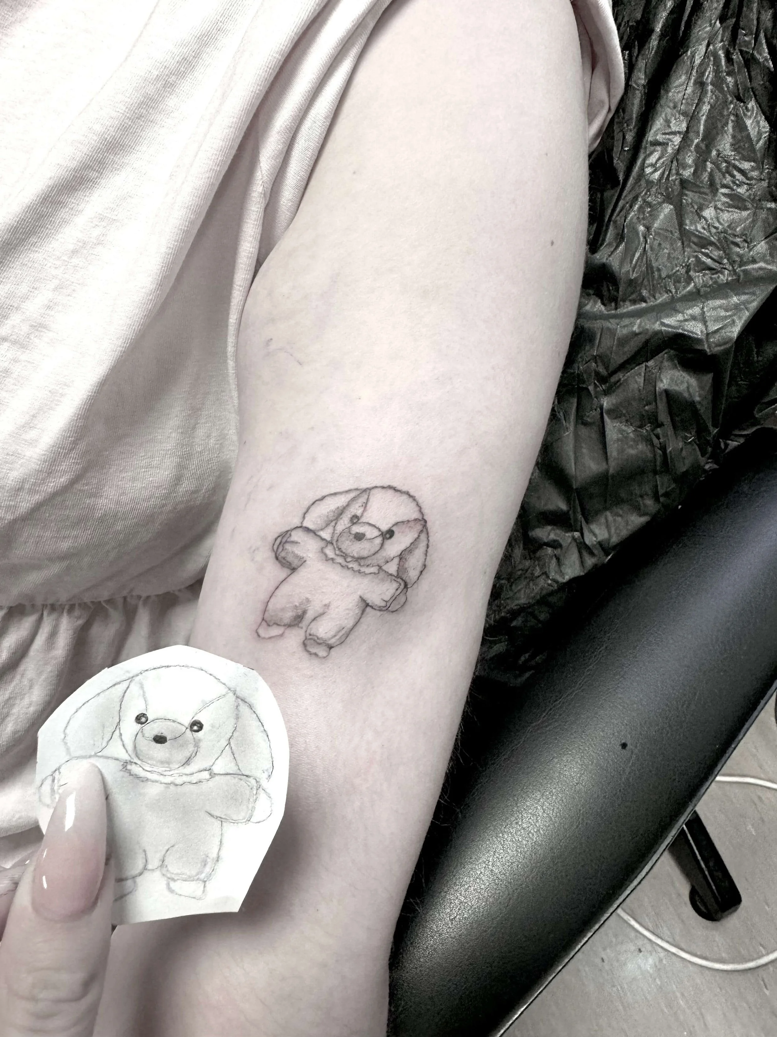 Delicate fine line upper arm tattoo of a cute fluffy stuffed animal dog or puppy toy, shown alongside its hand-drawn stencil reference. Soft minimal linework with gentle shading. Siren Tattoo Studio, Lucerne, Switzerland.