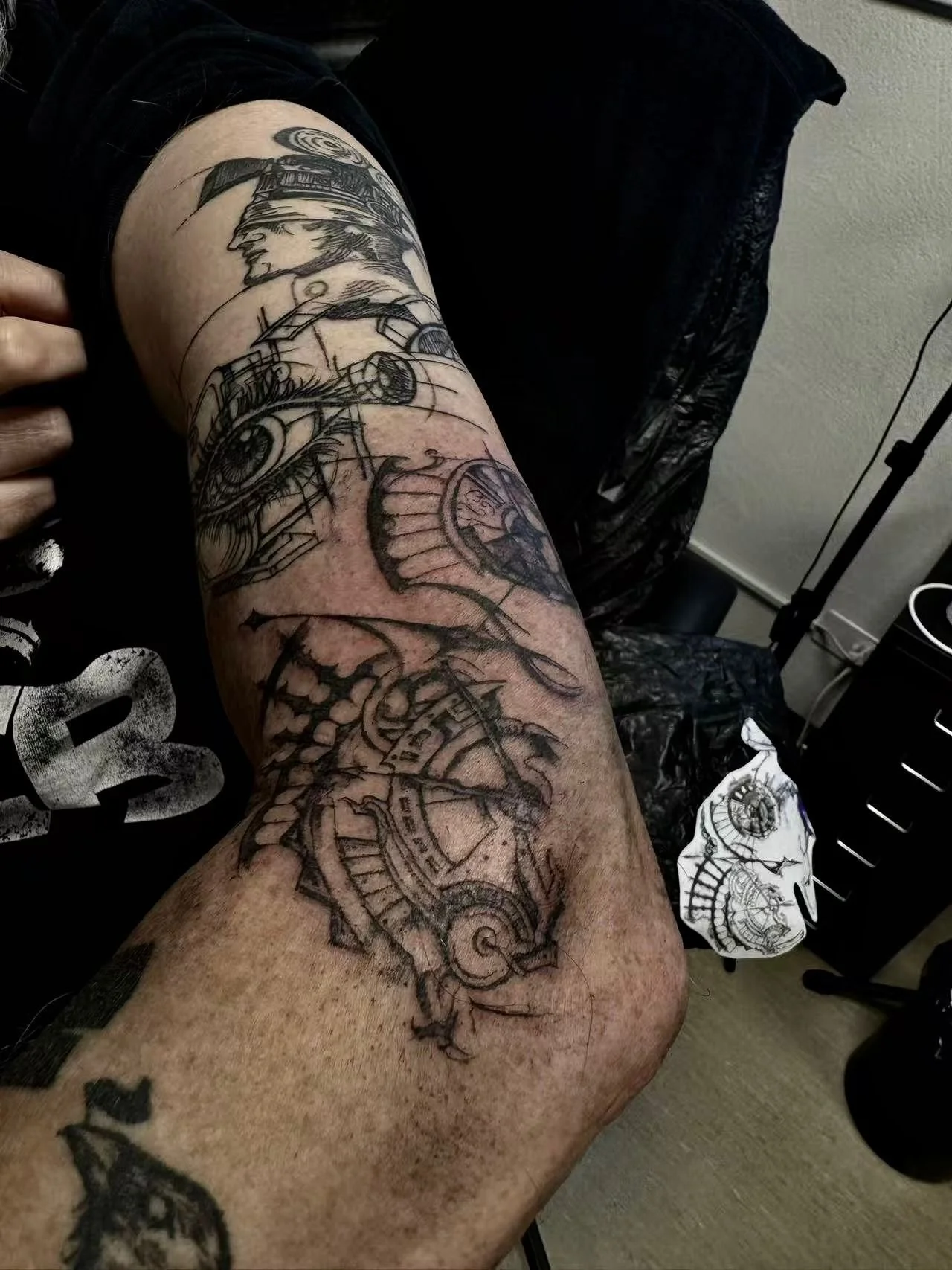 Fine line blackwork illustrative sleeve tattoo on the upper arm and forearm featuring a surrealist manga-style mechanical head portrait with camera reels at the shoulder, a detailed compass rose in the centre, and an ornate antique clock or astrolabe