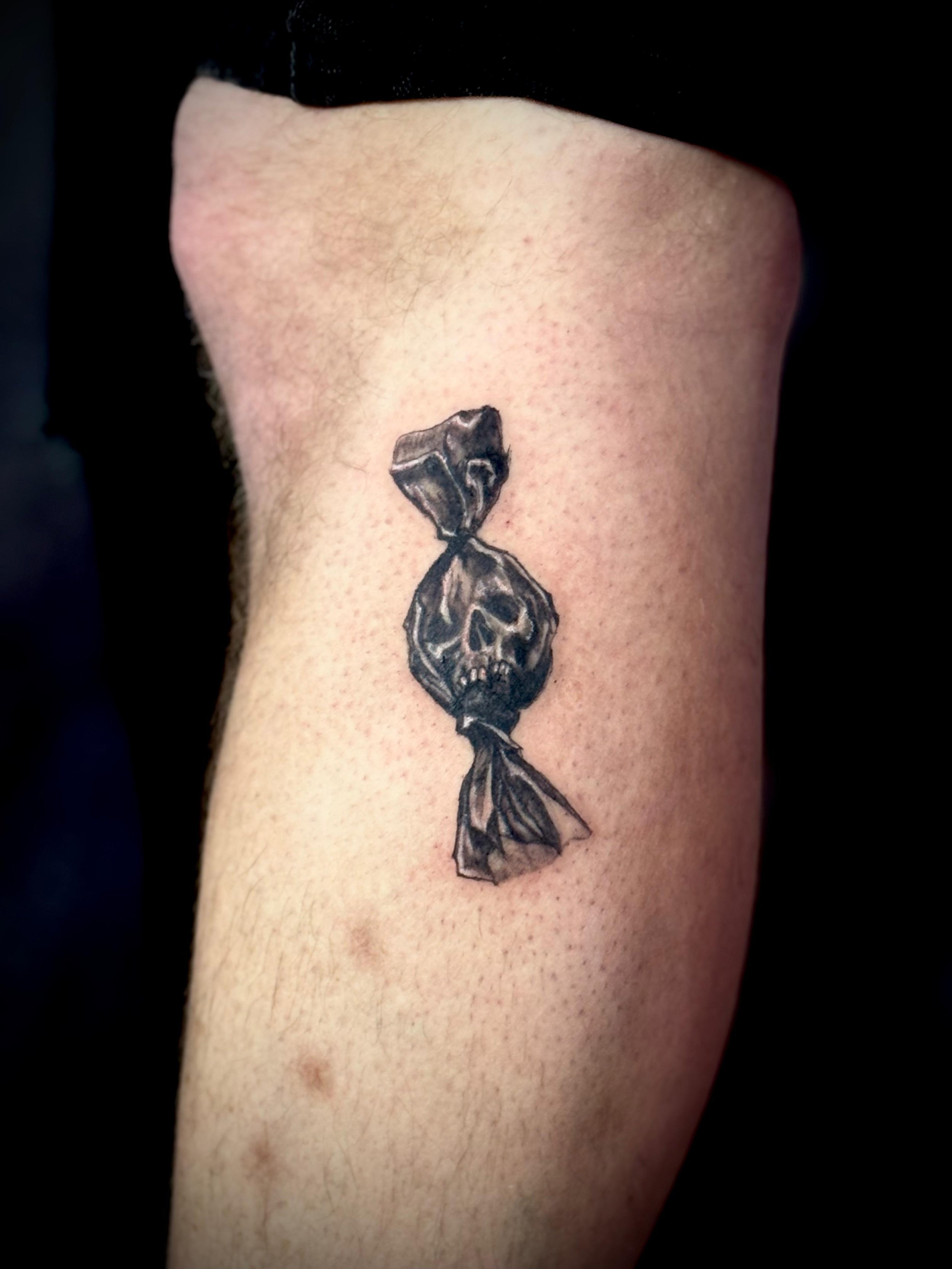 Small fine-line skull candy tattoo on knee — dark illustrative wrapped sweet with skull face in black and grey by Siren Tattoo Lucerne