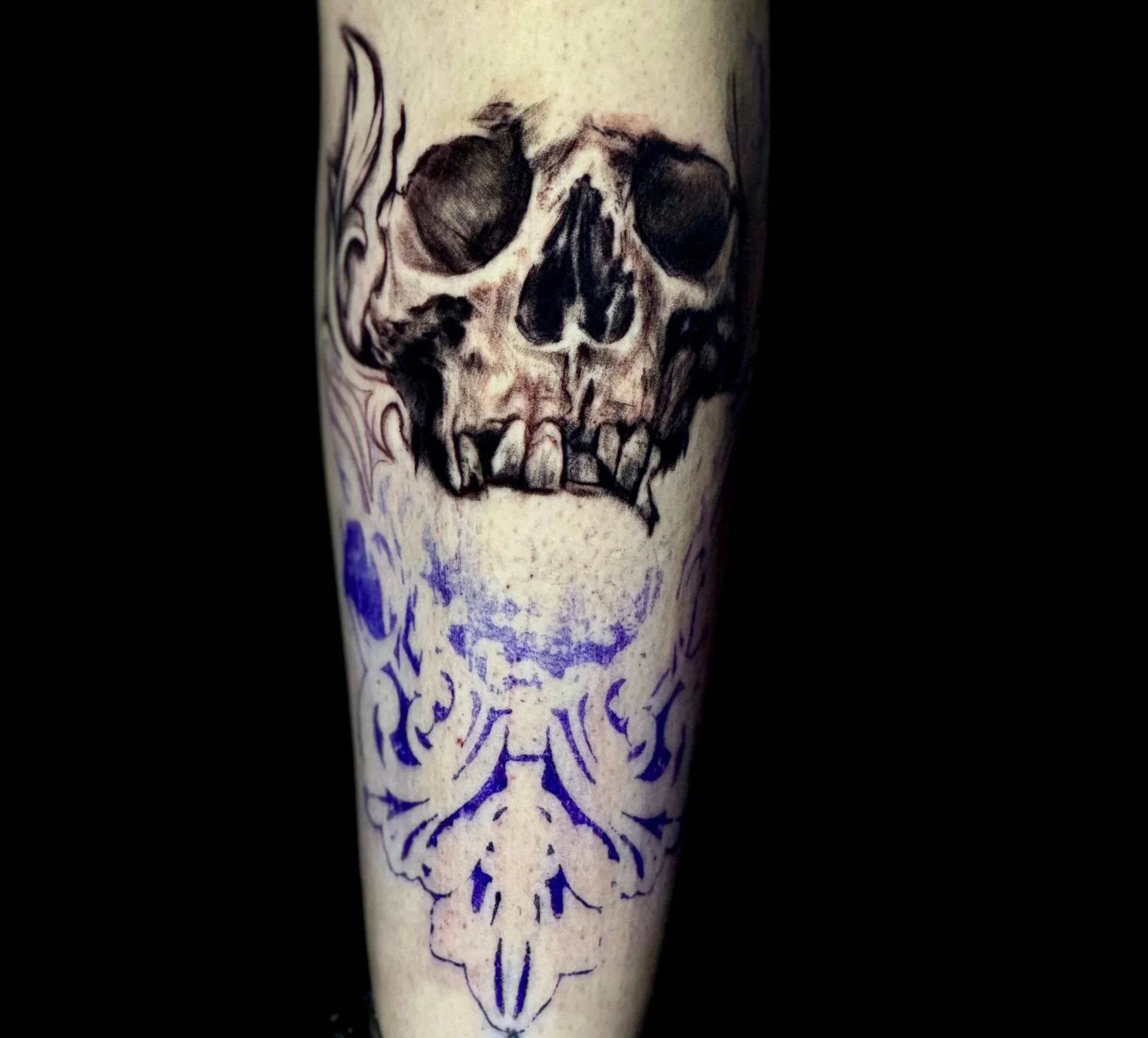 Close-up of a black and grey realism lower leg tattoo showing a detailed skull mid-session, with baroque ornament stencil marks still visible in blue below. Existing decorative scroll tattoo visible above. Siren Tattoo Studio, Lucerne, Switzerland.