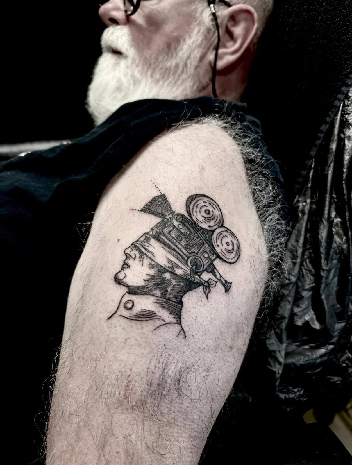 Fine-line illustrative shoulder tattoo — surrealist manga-style mechanical head portrait with camera reels in blackwork by Siren Tattoo Lucerne