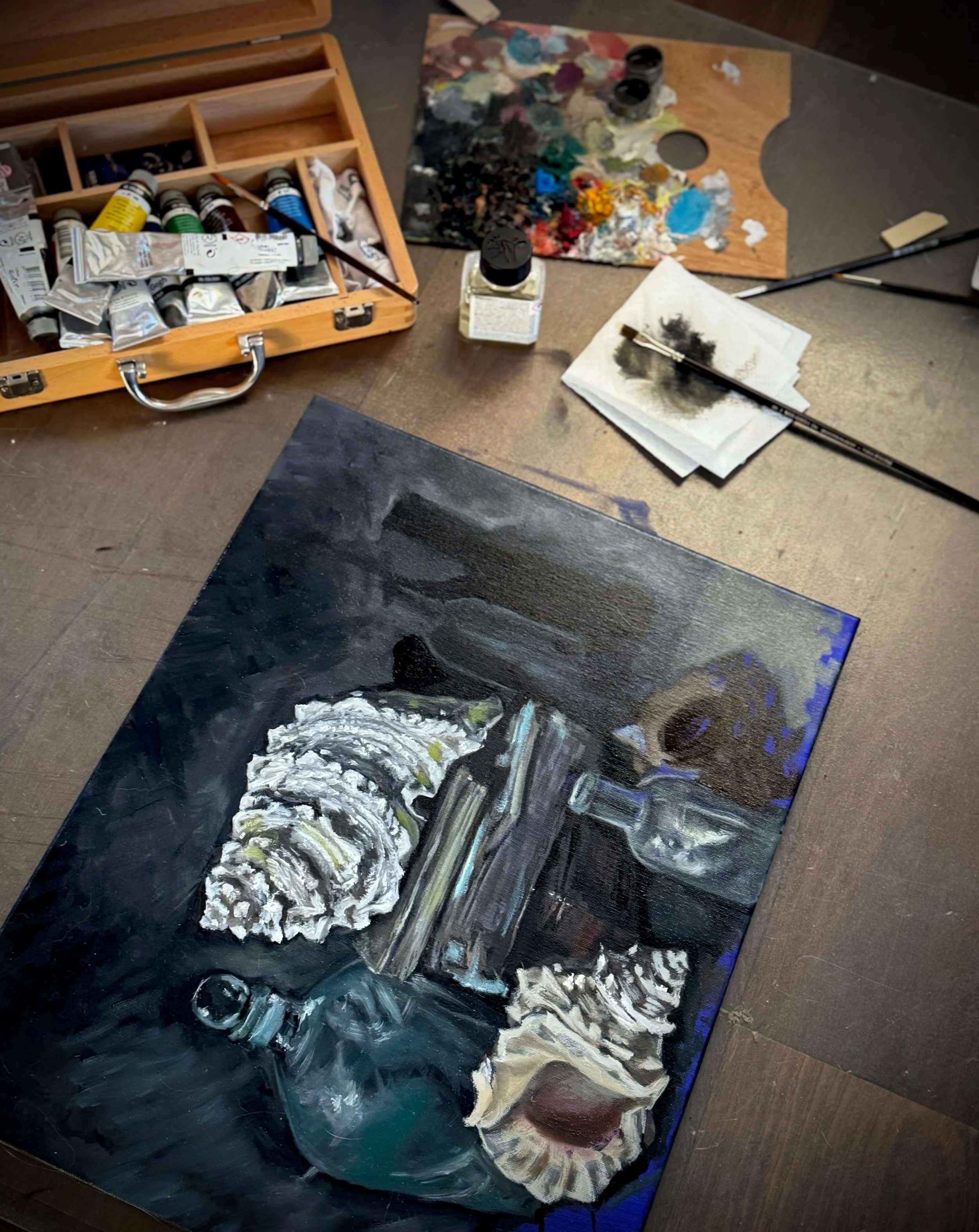 Completed dark still life oil painting on canvas showing seashells, a glass bottle, and stacked books arranged against a rich black and blue background, placed on a studio worktable alongside paint tubes, a wooden palette, and brushes. Siren Tattoo S