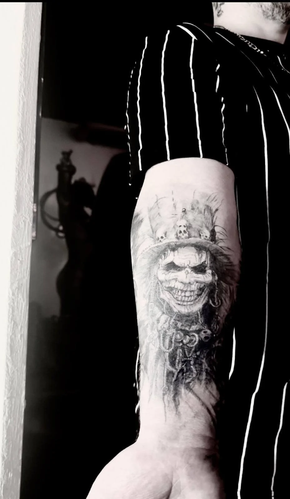 Black and grey forearm tattoo of a grinning skull figure wearing a feathered top hat decorated with small skulls, surrounded by dark ornamental details and flowing dark elements. Monochrome photograph. Siren Tattoo Studio, Lucerne, Switzerland.