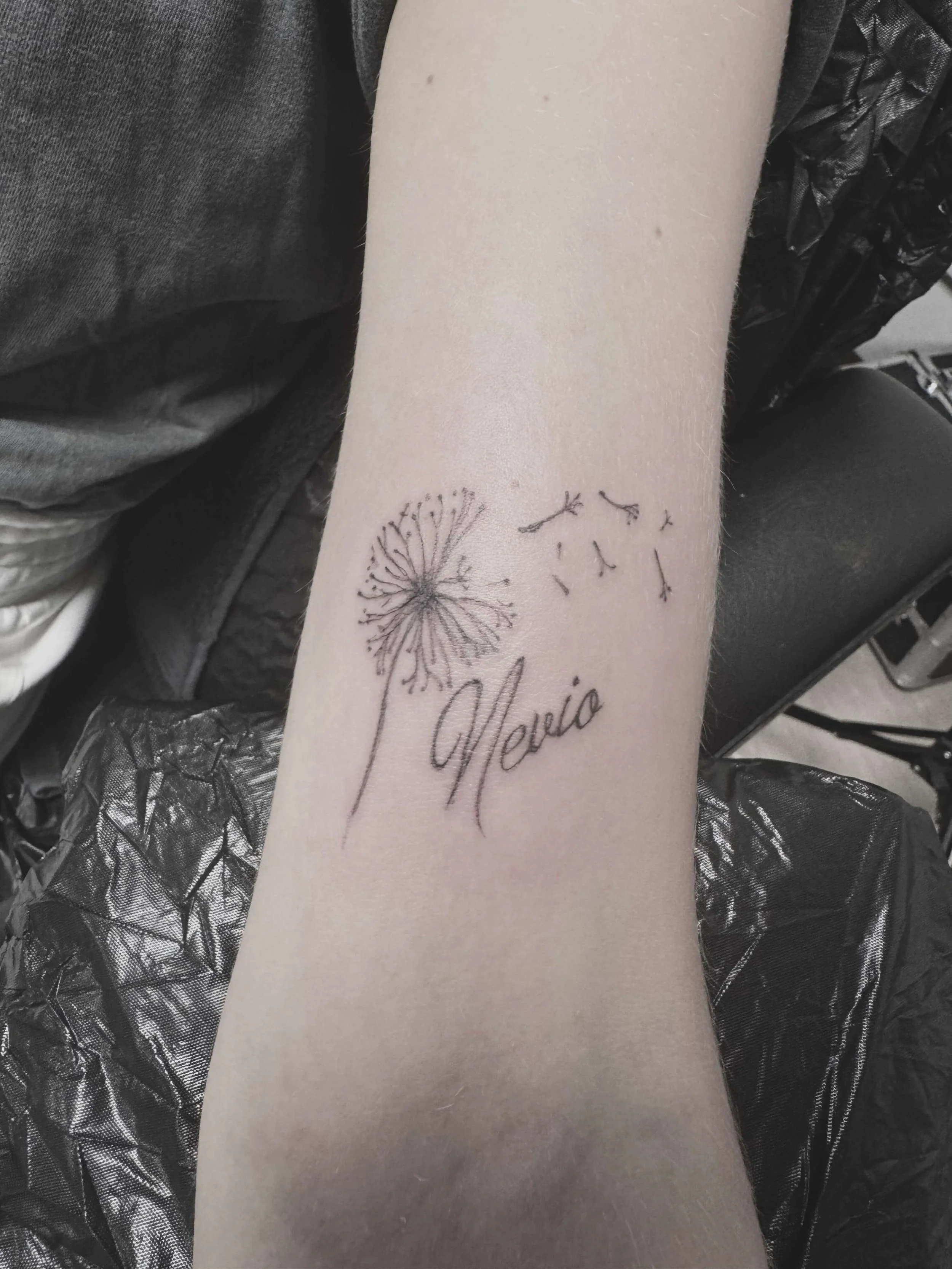 Fine-line dandelion and name tattoo on wrist — delicate botanical with cursive script lettering "Nevio" in single needle black by Siren Tattoo, Lucerne