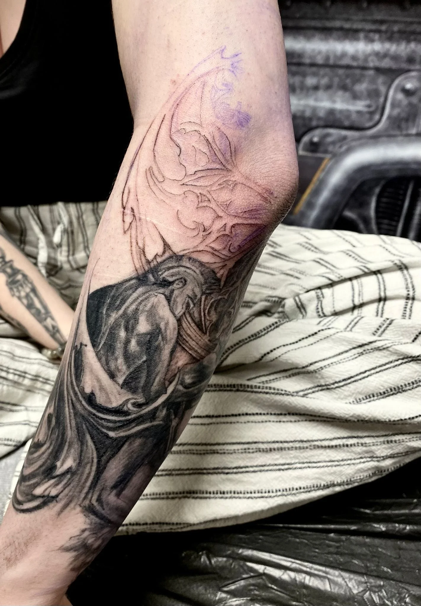 Work-in-progress black and grey realism forearm and elbow tattoo featuring a powerfully shaded dark angel or fallen figure — a muscular draped body with large wings spreading upward toward the elbow, rendered in deep blacks and soft grey gradients. W