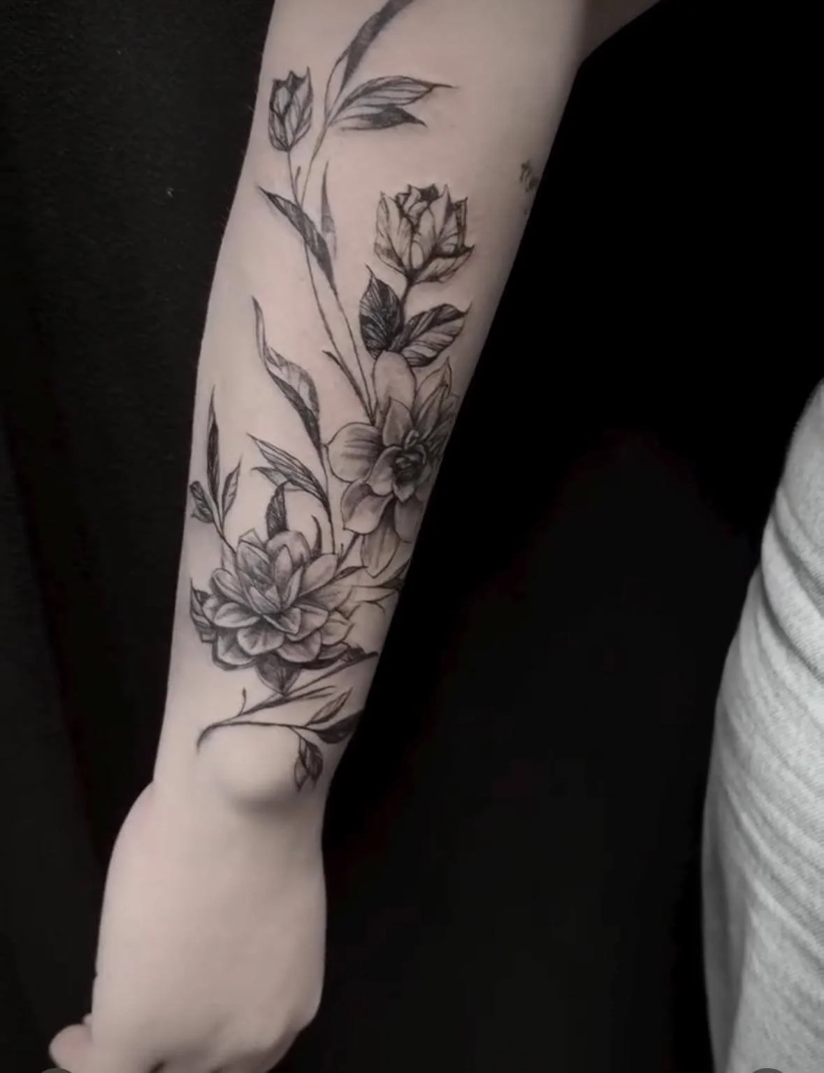Elegant black and grey botanical tattoo covering the forearm with a flowing arrangement of peonies, tulip buds, and leaves rendered in fine linework and soft shading. Delicate floral sleeve design. Siren Tattoo Studio, Lucerne, Switzerland.
