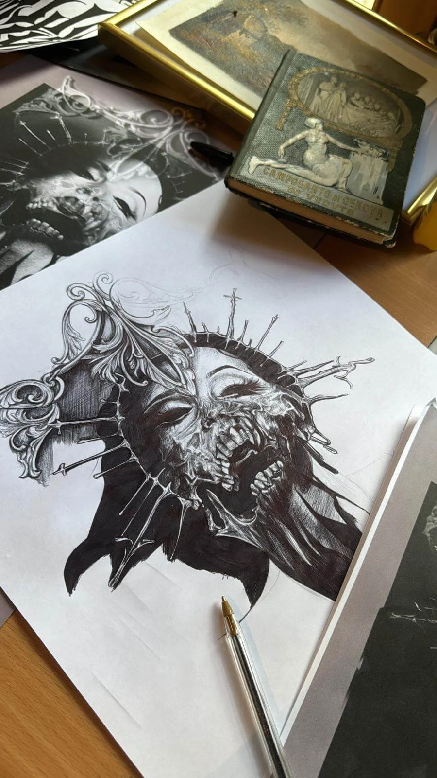 Original ink drawing on paper — screaming skull with thorn crown, baroque scrollwork and daggers, studio desk with vintage book and reference prints at Siren Tattoo Lucerne