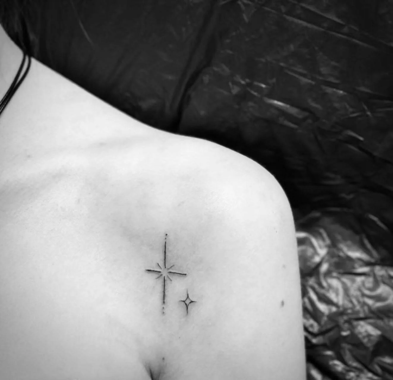 Minimal fine-line star tattoo on collarbone — two four-pointed sparkle stars in single needle black on chest by Siren Tattoo, Lucerne