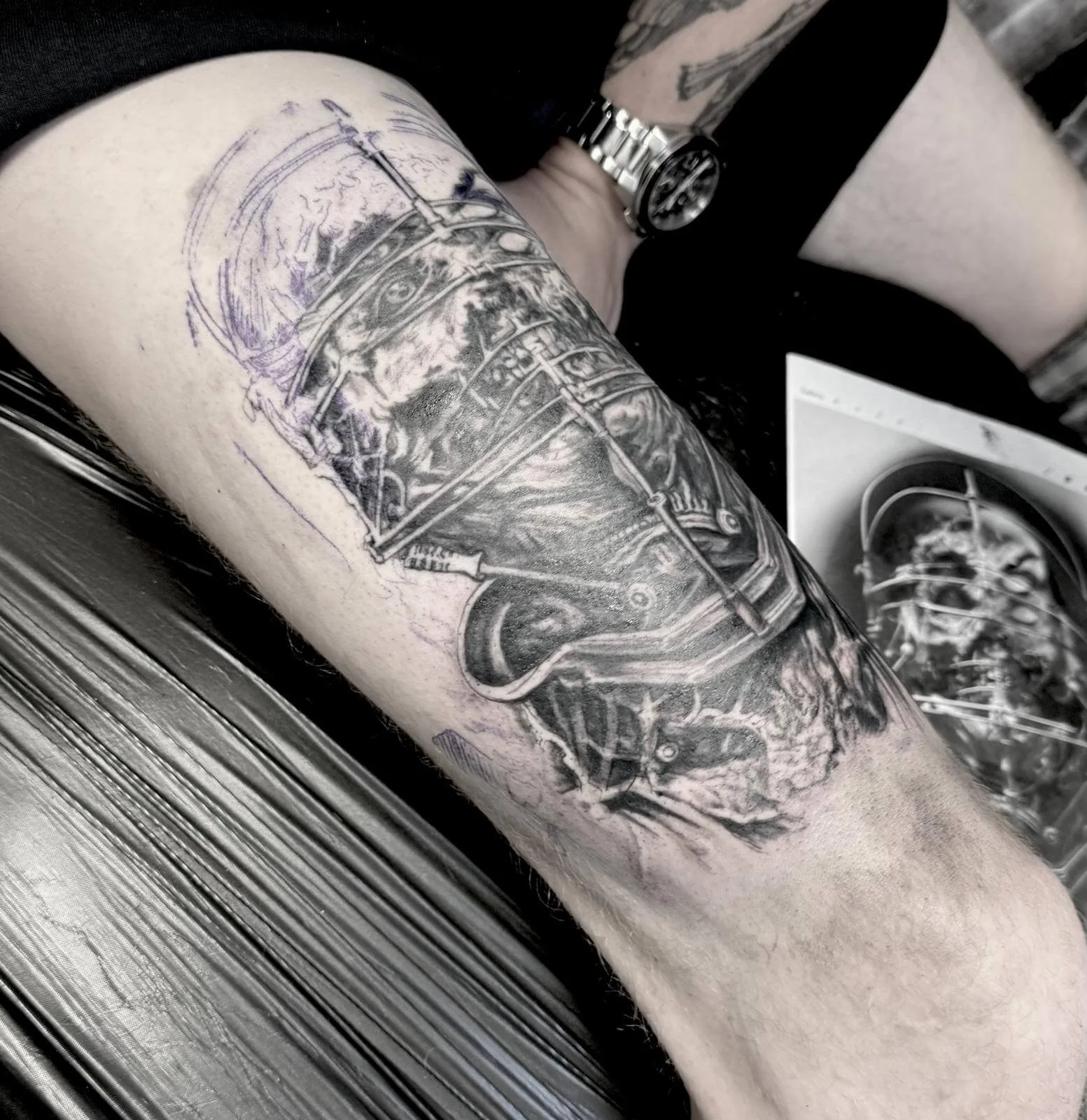 Completed black and grey realism tattoo on the thigh depicting a disturbing figure with a cage-like metal headpiece constraining a decayed or tortured face, rendered in hyperrealistic detail. Reference drawing shown alongside. Siren Tattoo Studio, Lu