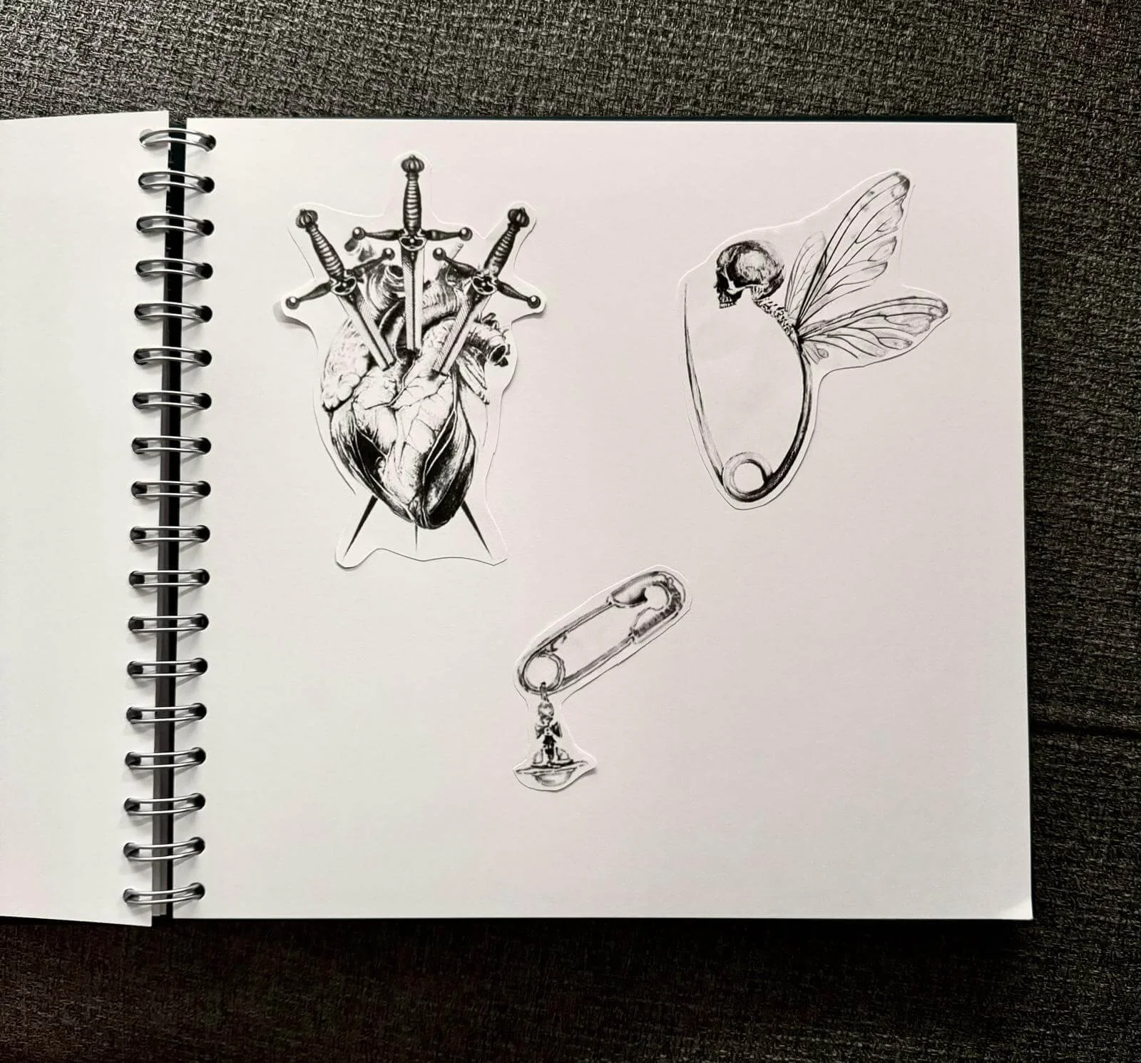 Sketchbook page with dark illustrative flash sticker designs — pierced anatomical heart with daggers, skull fairy on safety pin, and hanging figure charm by Siren Tattoo Lucerne