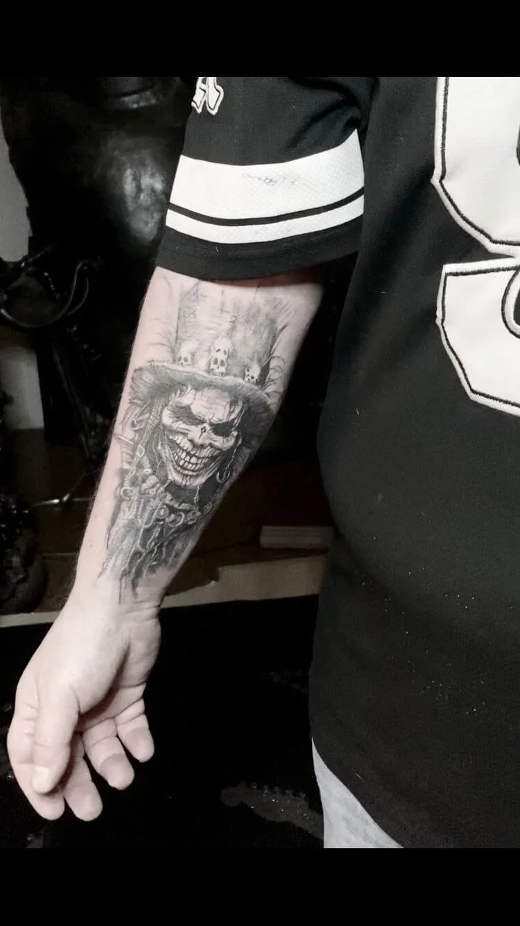 Black and grey forearm tattoo of a grinning skull figure wearing a feathered top hat decorated with small skulls, surrounded by dark ornamental details and flowing dark elements. Monochrome photograph. Siren Tattoo Studio, Lucerne, Switzerland