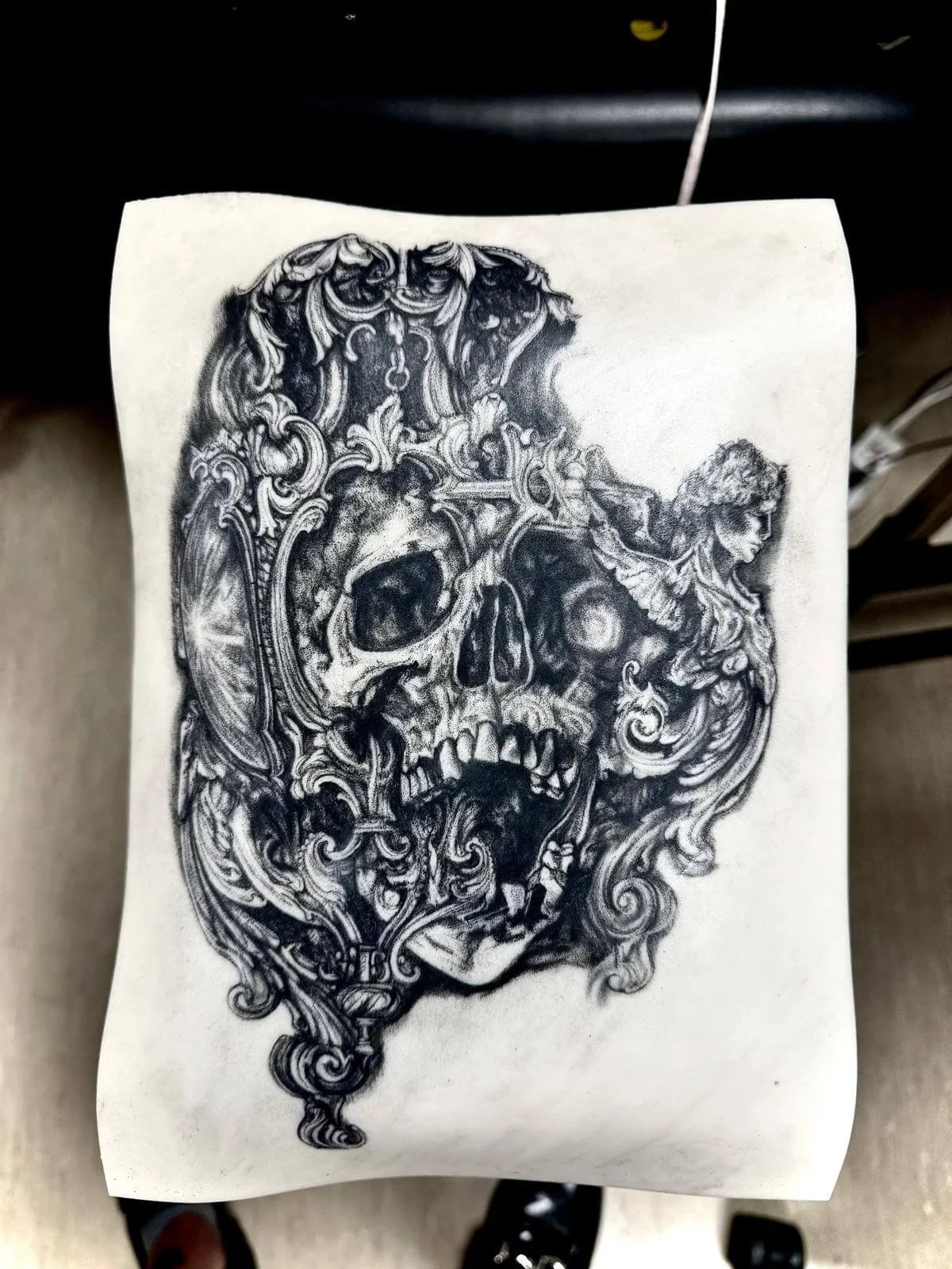Practice piece on fake skin — hyperdetailed baroque skull with ornate frame, scrollwork and cherub figure by Siren Tattoo Lucerne