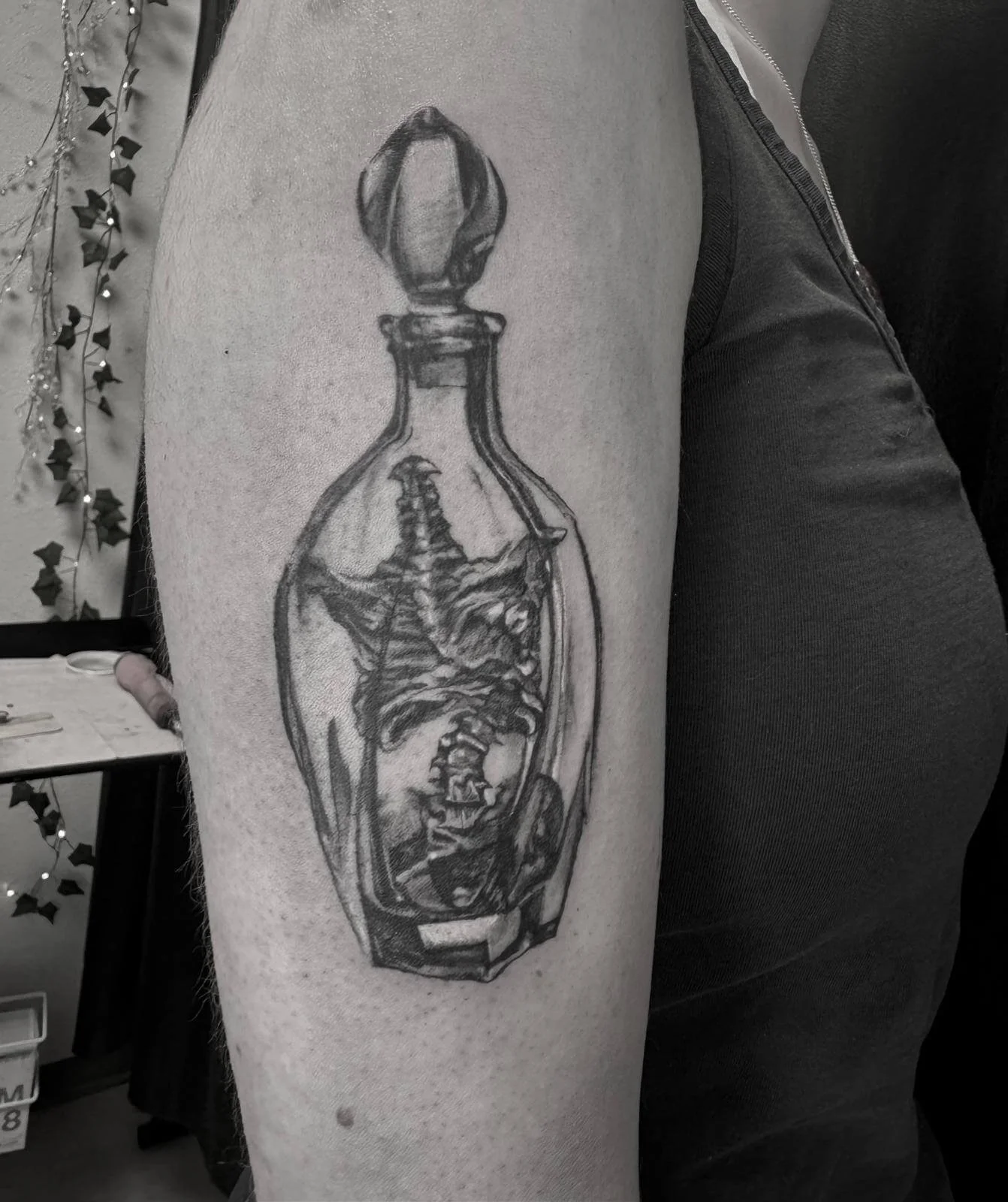 Surrealist blackwork bottle tattoo on upper arm — skeletal creature and spine contained within glass decanter, fresh linework by Siren Tattoo Lucerne