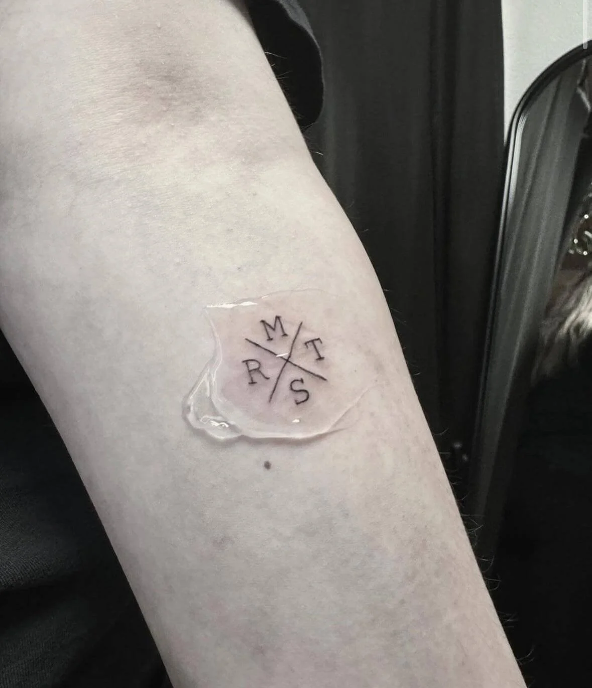 Minimalist fine line tattoo on the inner forearm showing four initials — M, T, R, S — arranged around a central cross in a compass-like layout, freshly done and covered with healing film. Siren Tattoo Studio, Lucerne, Switzerland.