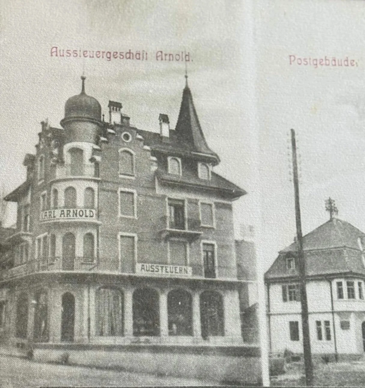 Framed vintage black and white postcard of the historic corner building housing Siren Tattoo Studio, captioned "Aussteuergeschäft Arnold" with an ornate onion dome tower and pointed turret. Lucerne, Switzerland.