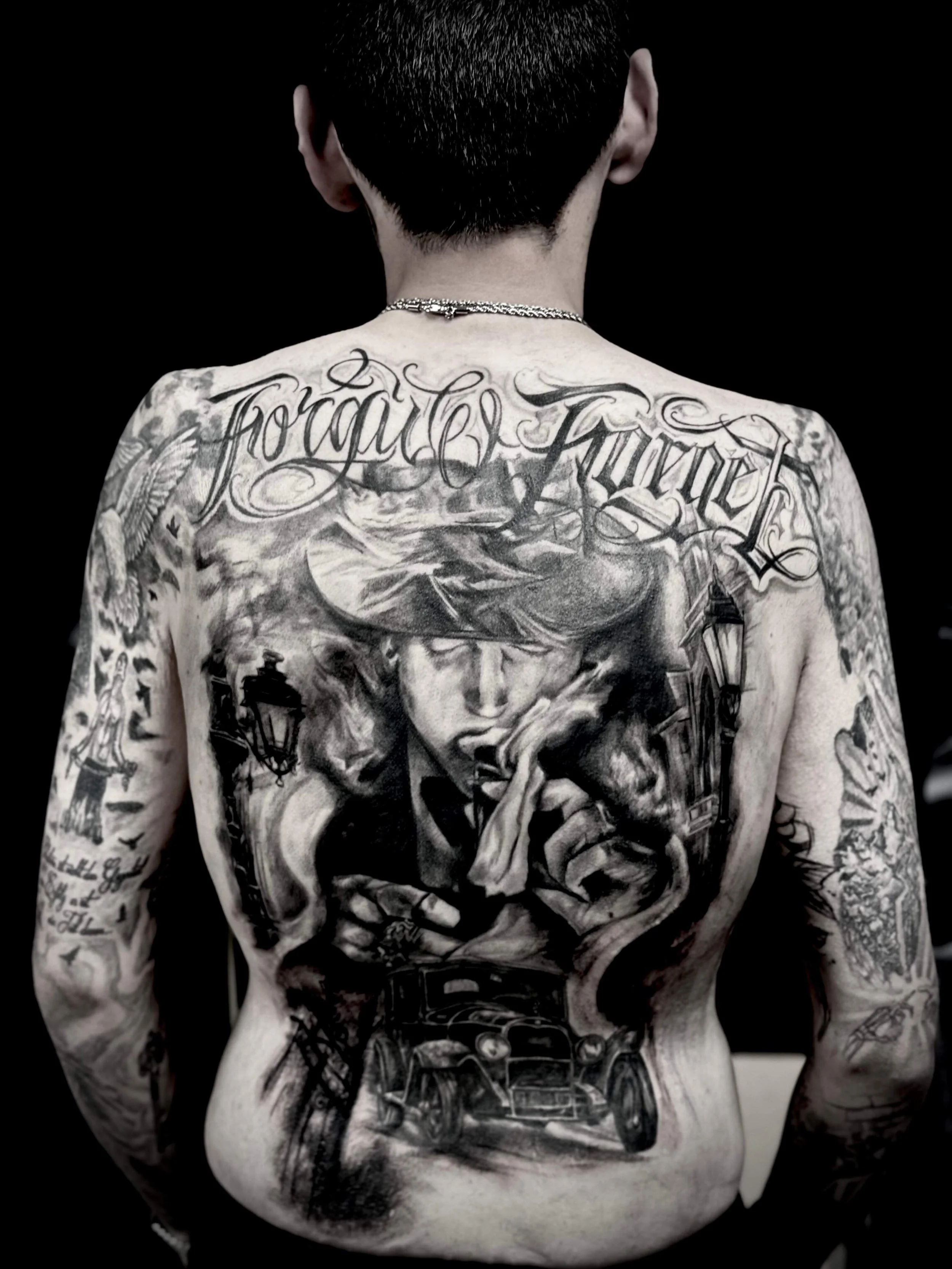 Full back tattoo in black and grey realism — "Forgive and Forget" script with cinematic portrait, skull, lantern and vintage car by Siren Tattoo Lucerne