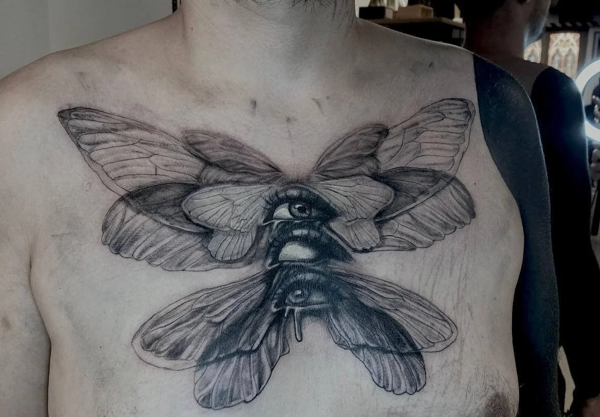 Black and grey surrealist chest tattoo depicting a large moth or butterfly with spread wings, its body replaced by a realistic human eye shedding a tear. Symmetrical placement across the sternum. Siren Tattoo Studio, Lucerne, Switzerland.