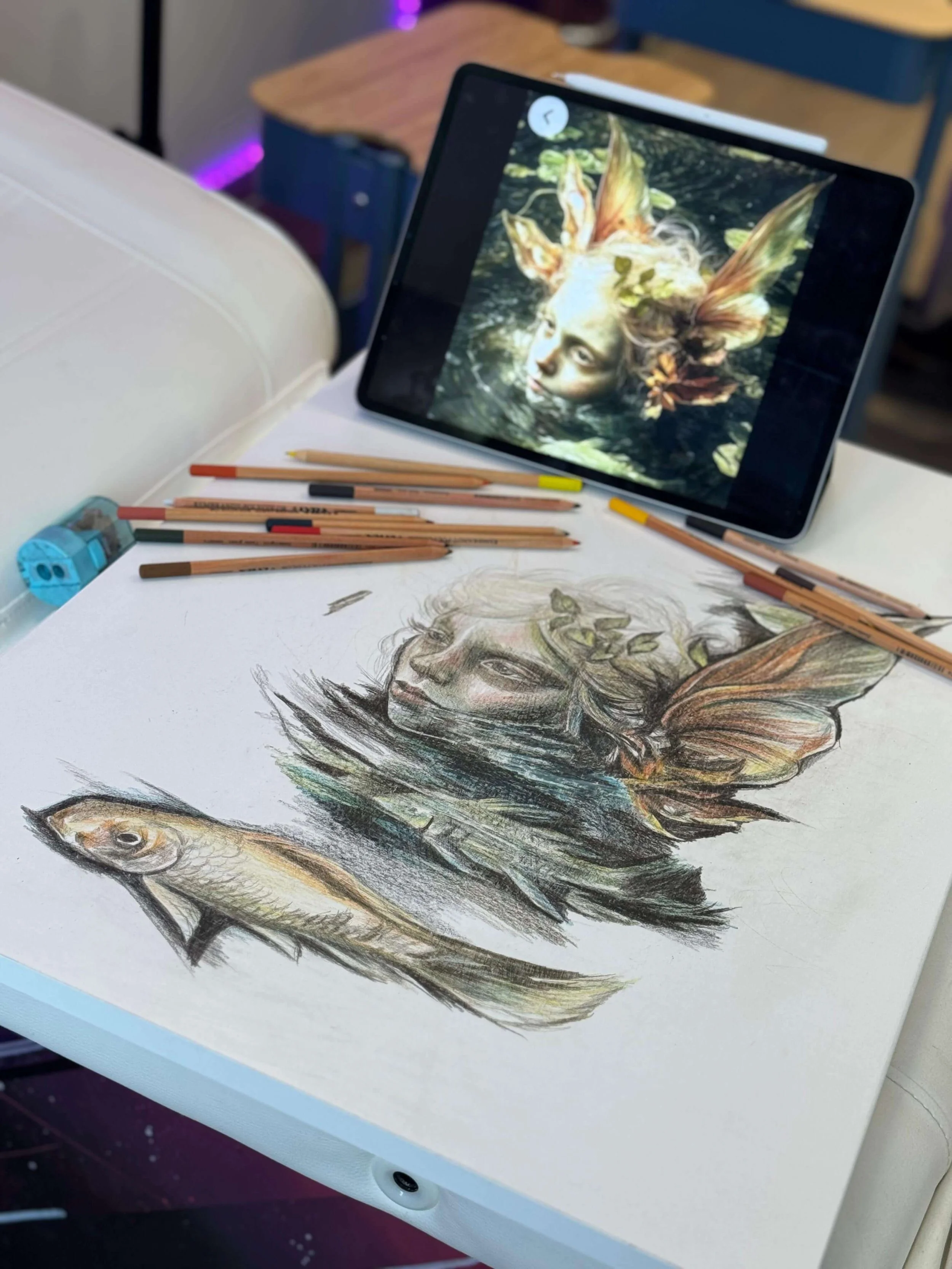 Tattoo artist's workspace showing a hand-drawn design in progress on paper — a fantasy nature spirit with a fish, butterfly wings, and leafy elements — alongside a reference image on a tablet screen. Coloured pencils and a sharpener visible on the dr