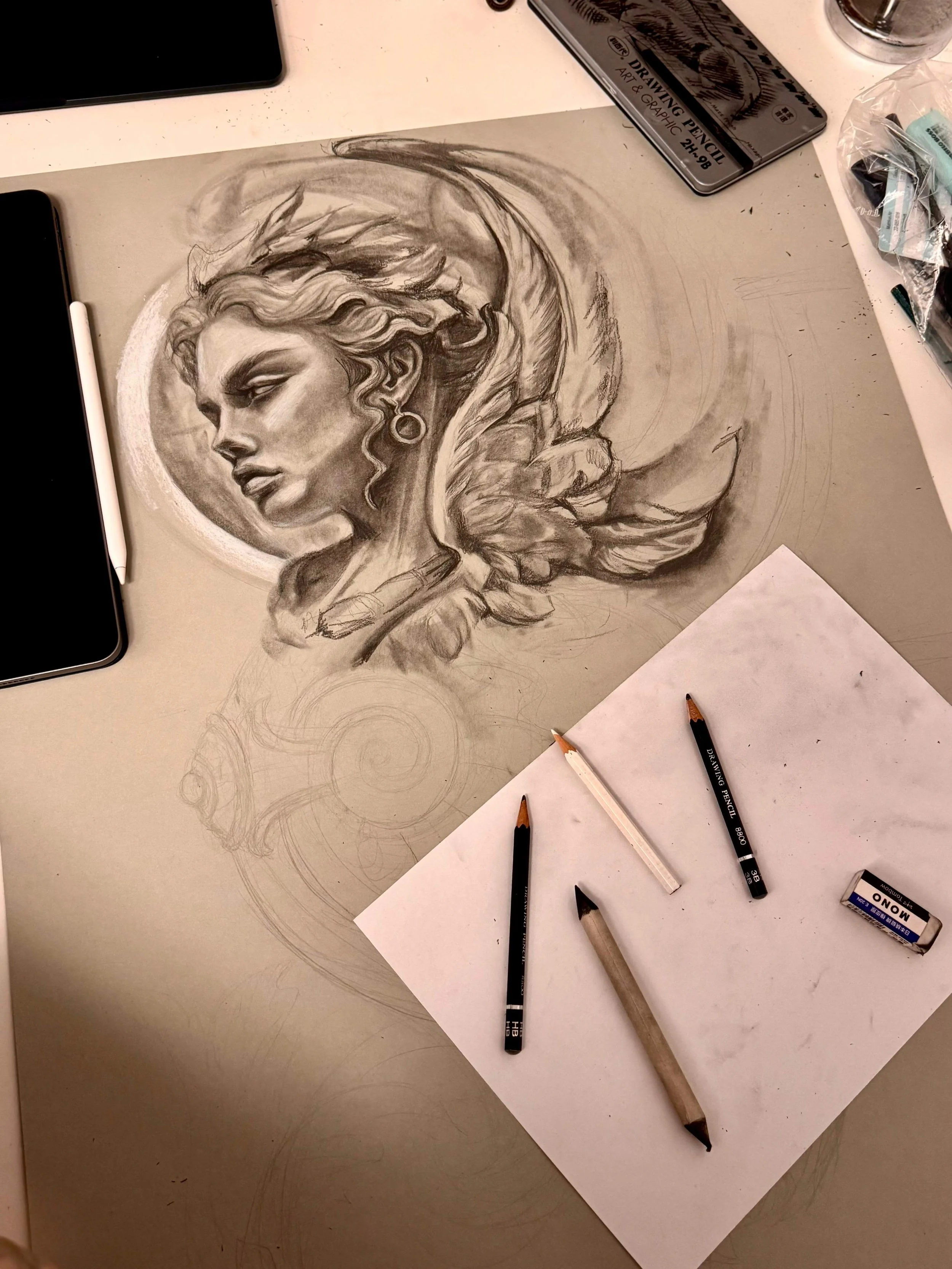 Original pencil drawing in progress on studio desk — winged female portrait with crescent moon and floral detail, tattoo design study by Siren Tattoo Lucerne