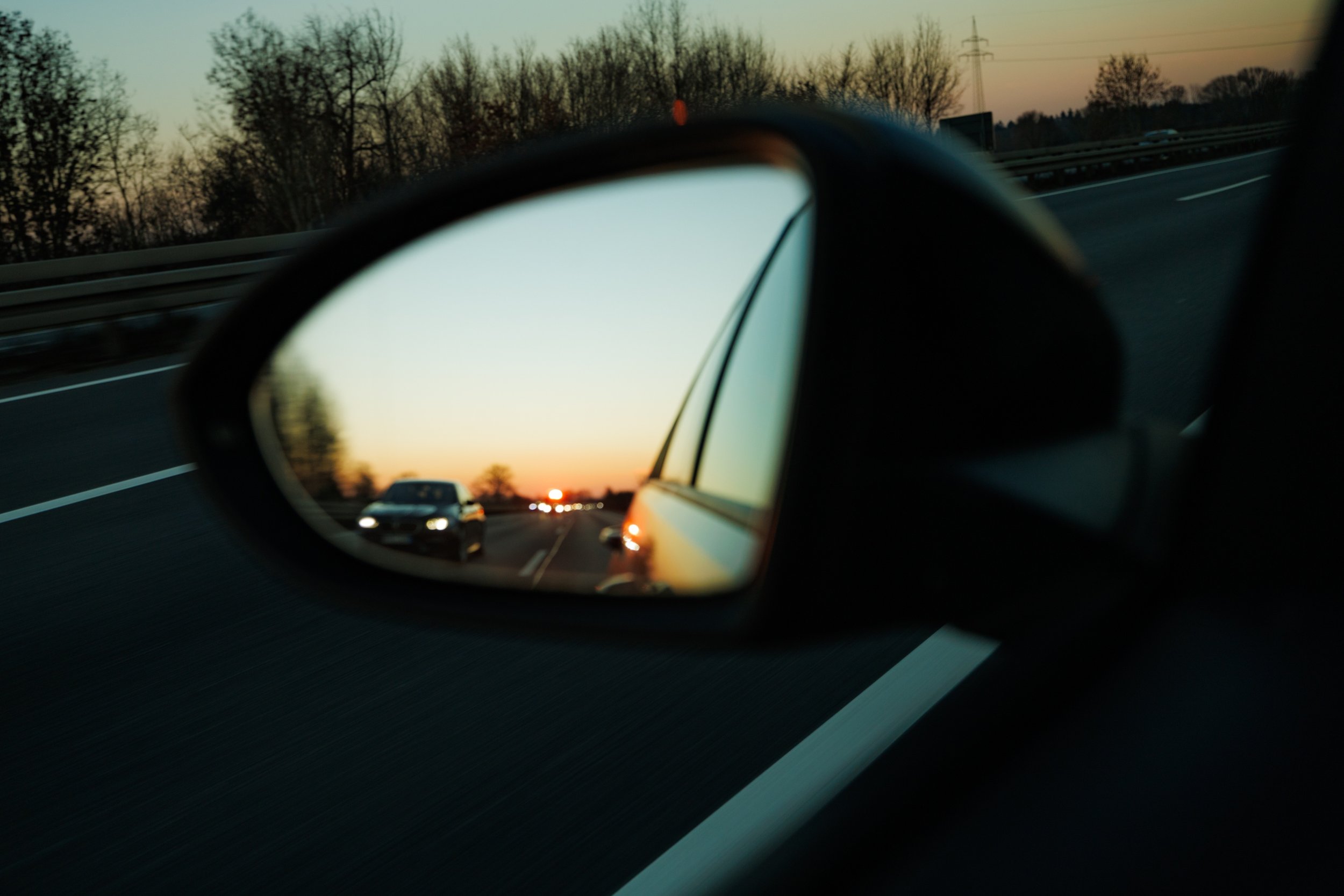 Blick in einen Rückspiegel während der Fahrt auf einer Autobahn bei Sonnenuntergang, mit anderen Autos und Bäumen im Hintergrund.