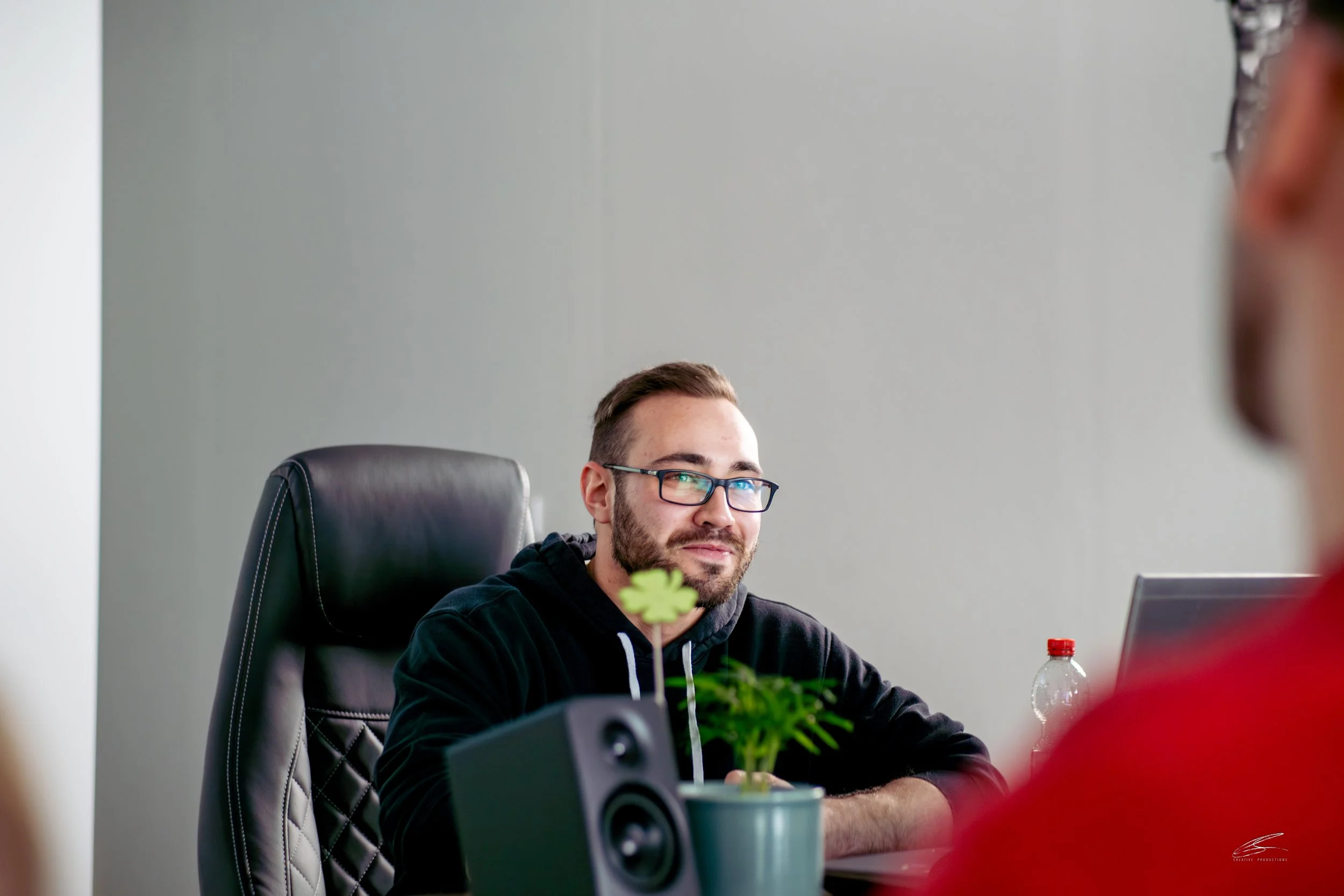 Ein Mann mit Brille und dunklem Hoodie sitzt an einem Tisch in einem Büro, vor ihm ein kleines Grünpflanzen, eine Wasserflasche und ein Laptop, im Vordergrund eine Person in rotem Shirt. Ein Lautsprecher steht auf dem Tisch.