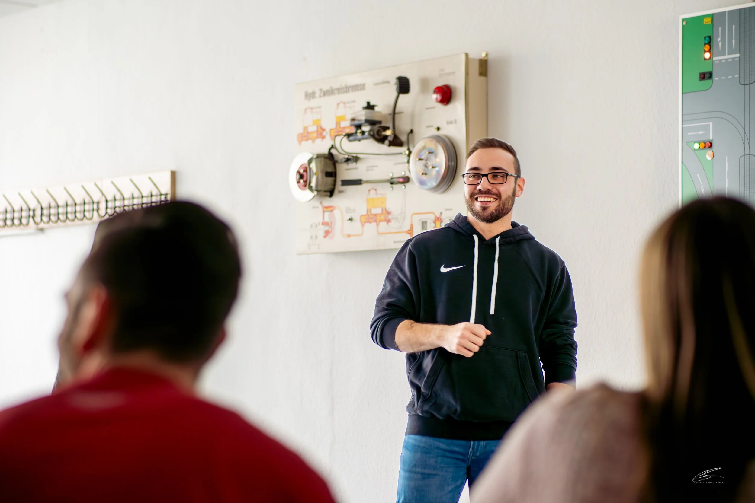Ein junger Mann mit Brille und dunklem Hoodie spricht vor einer Gruppe während eines Unterrichts. Im Hintergrund sind technische Poster an der Wand.