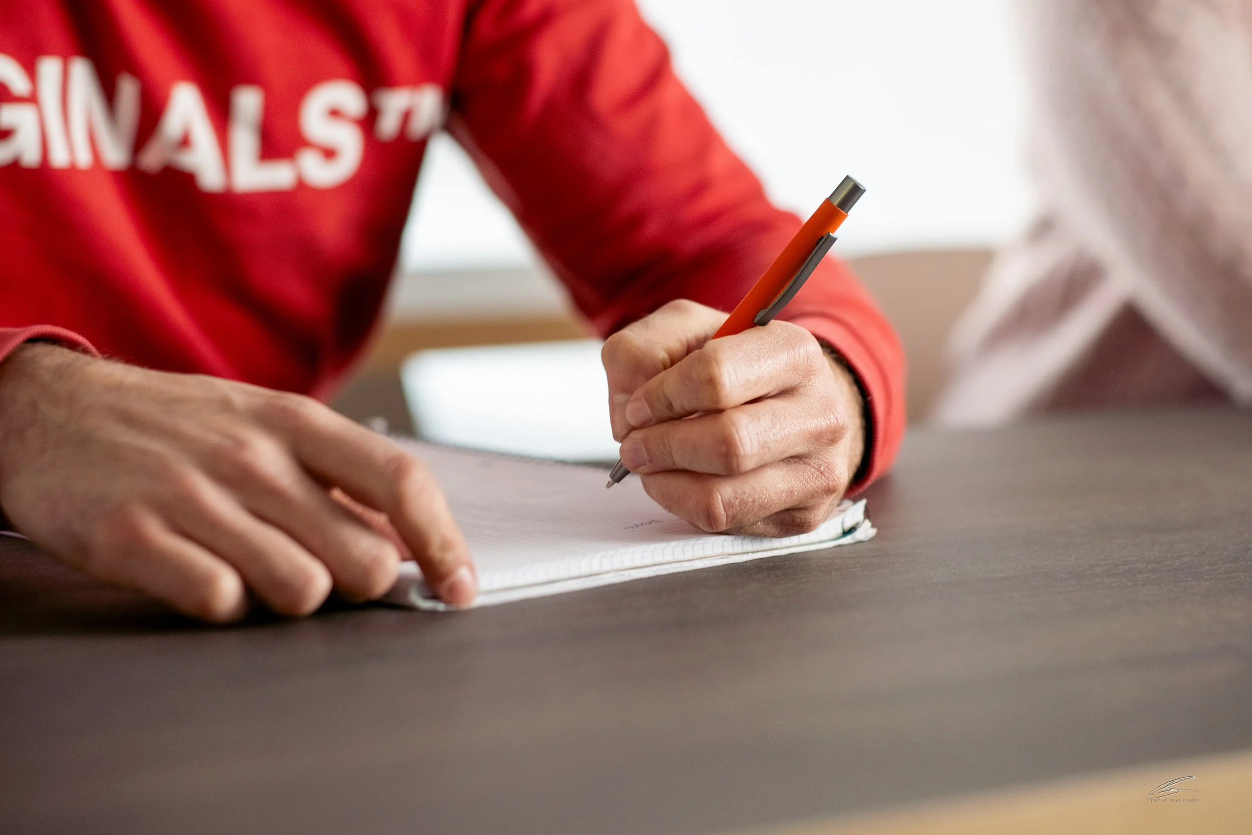 Person schreibt auf einem Notizbuch mit einem Stift, während sie an einem Tisch sitzt.