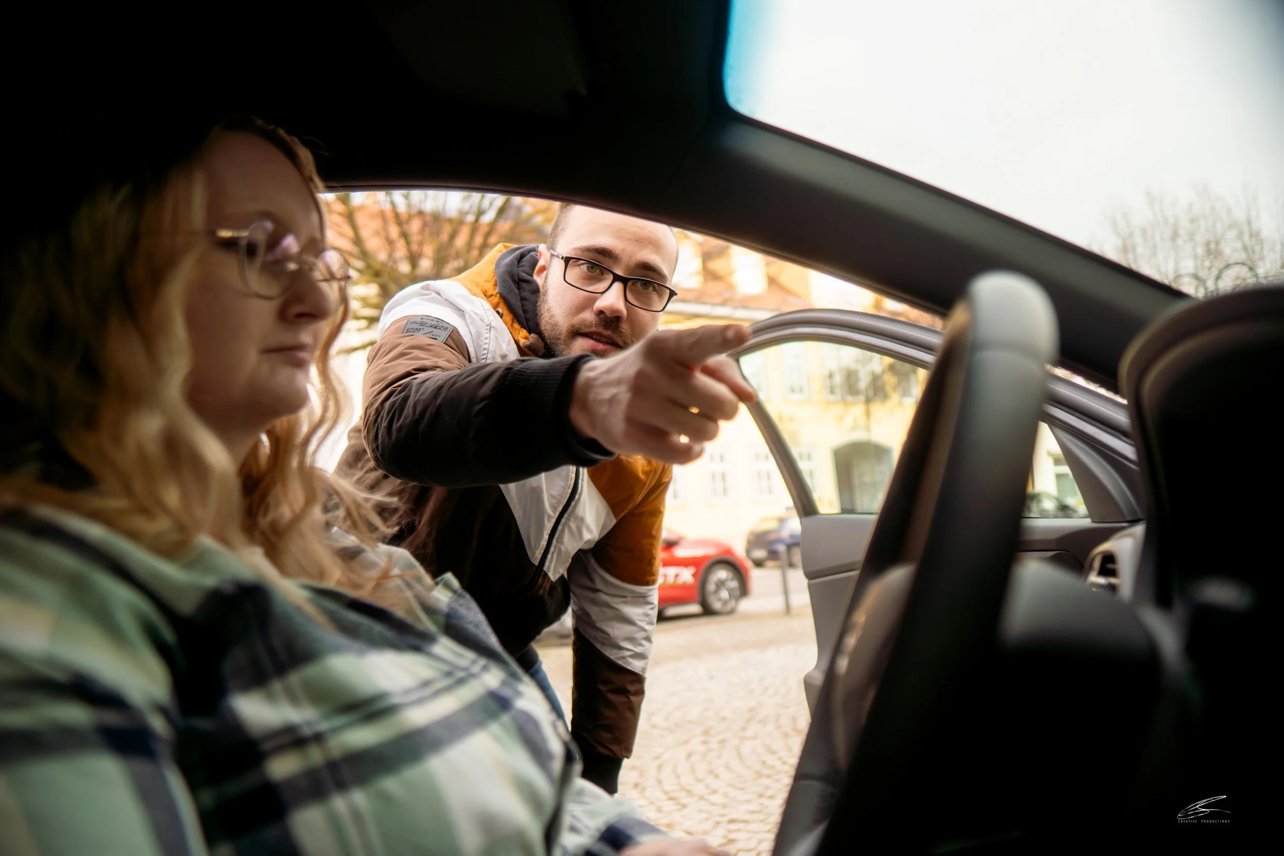 Ein Mann mit Brille zeigt auf die offene Tür eines Autos, während eine Frau mit Brille und kariertem Hemd im Inneren des Autos sitzt. Im Hintergrund sind Häuser und ein rotes Auto auf der Straße zu sehen.