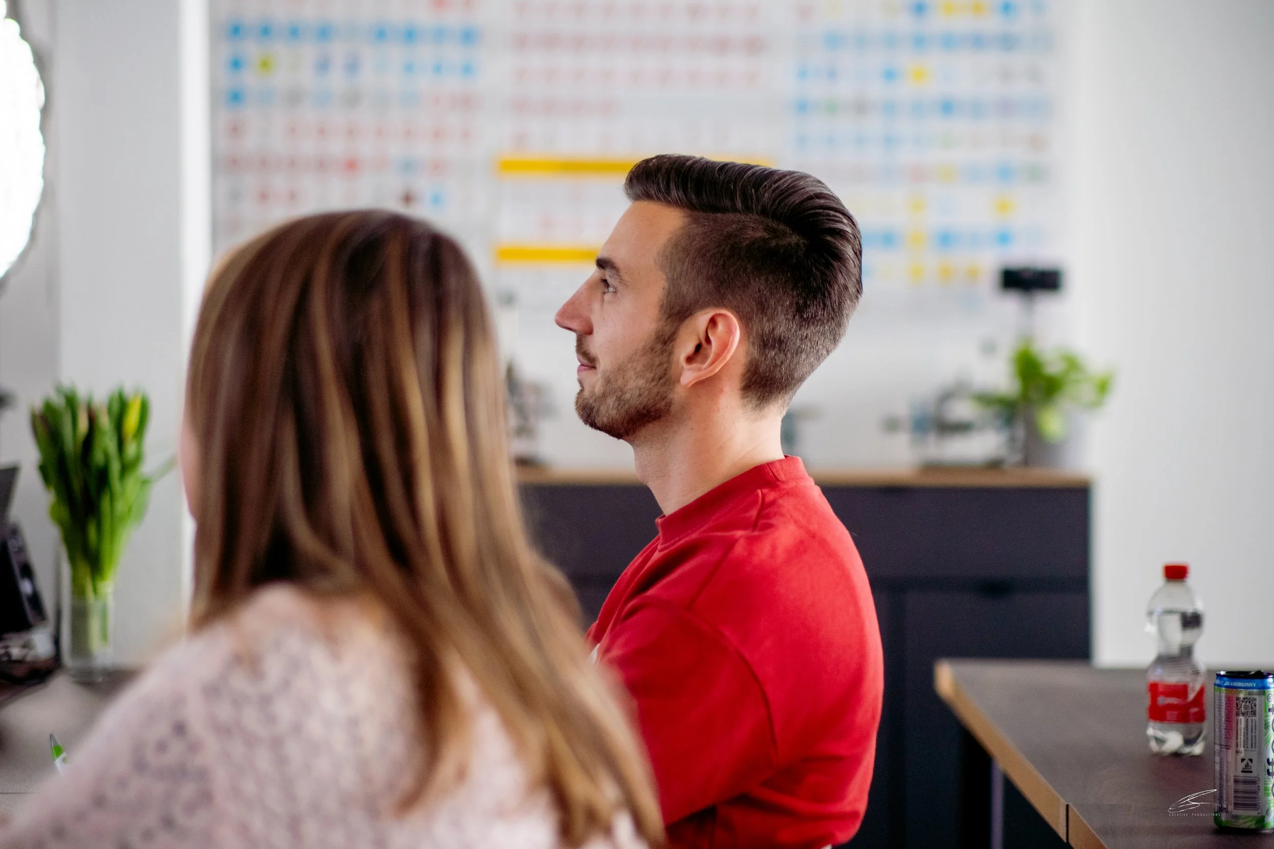 Zwei Menschen in einem Büro oder Konferenzraum, die sich gegenüber sitzen, während im Hintergrund eine große Tafel mit Farben und Markierungen zu sehen ist.