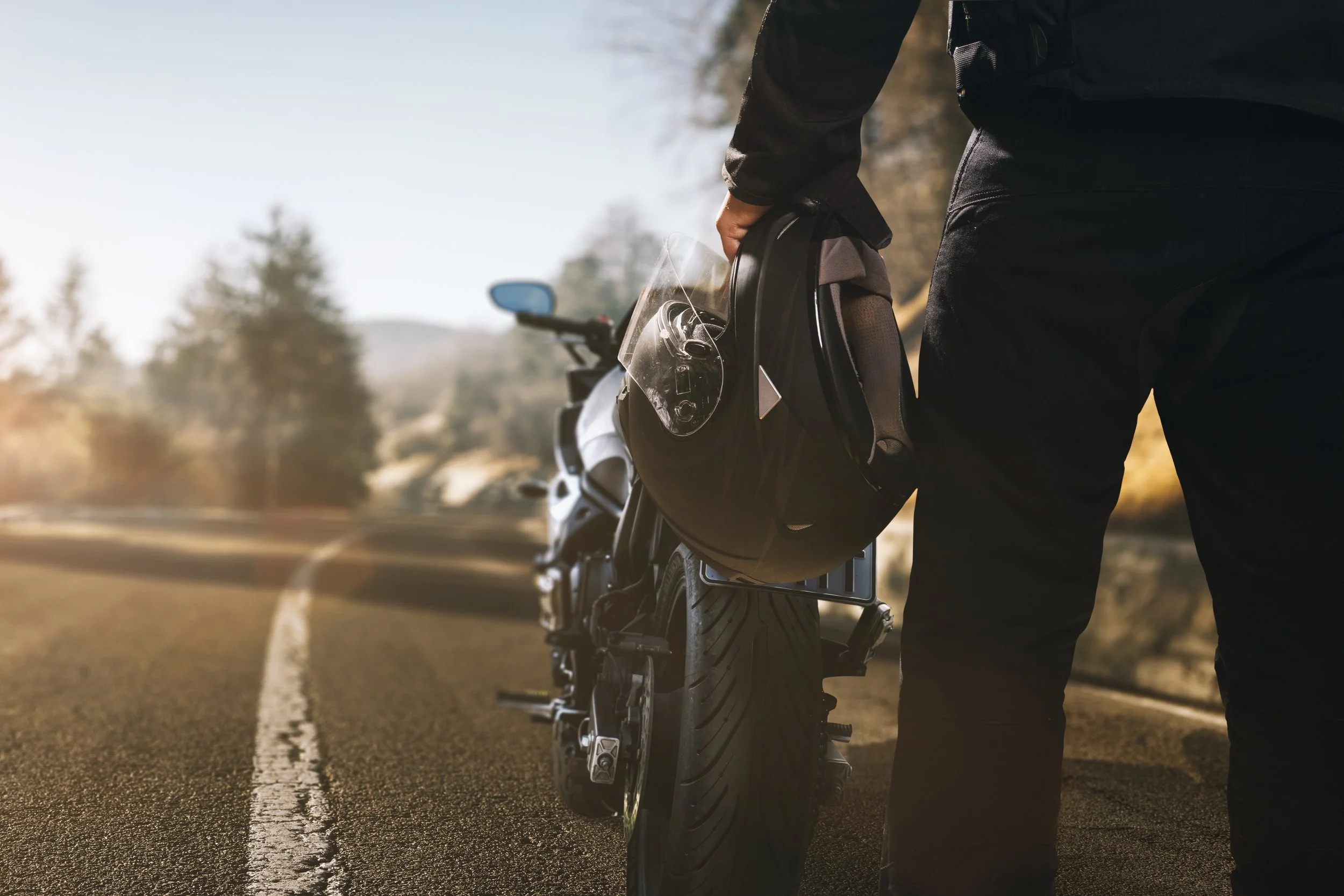 Person mit schwarzer Kleidung steht neben einem Motorrad auf einer Straße, hält einen schwarzen Helm in der Hand, im Hintergrund Bäume und eine bewölkte Landschaft bei Sonnenaufgang oder Sonnenuntergang.